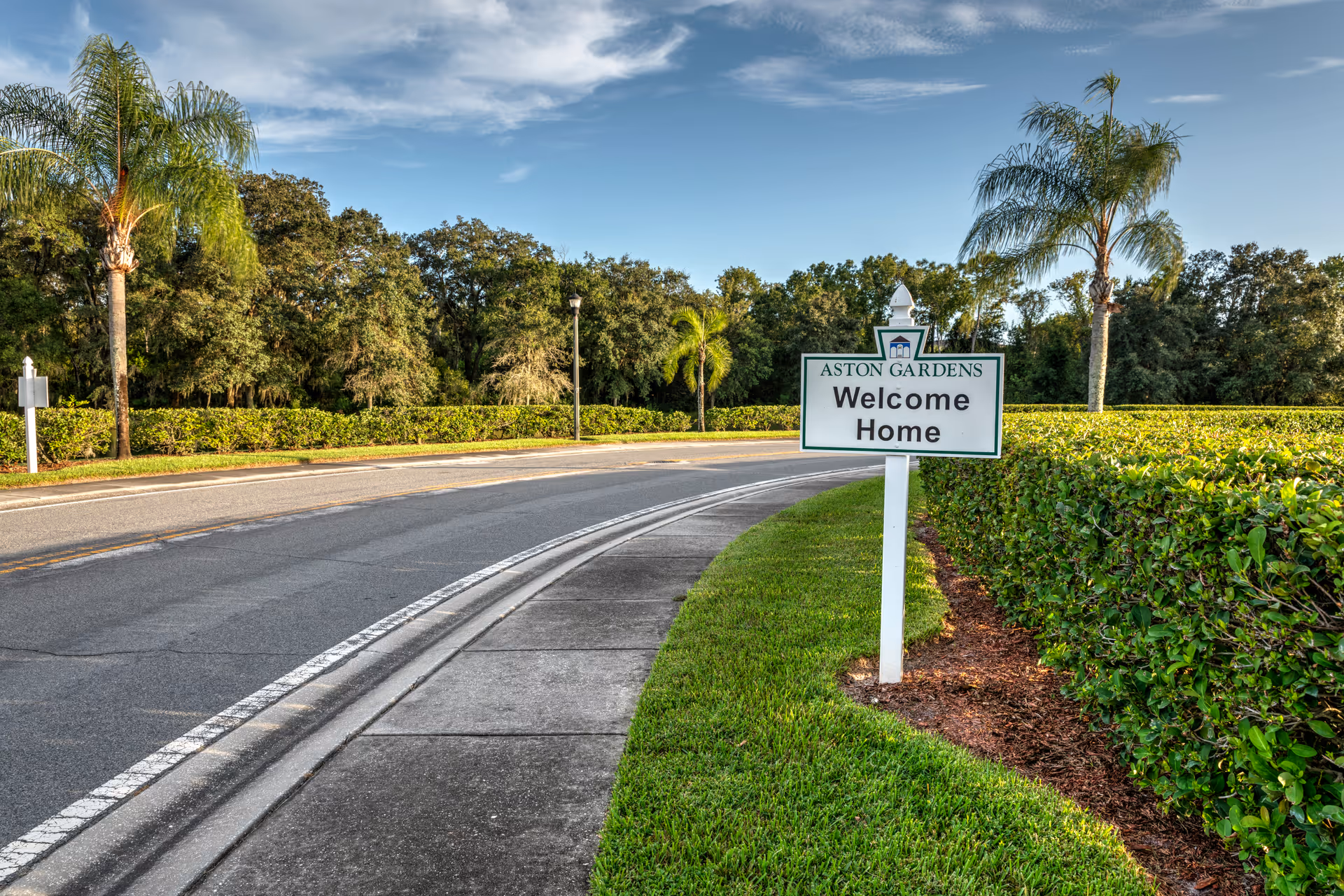A curved road with a sidewalk and neatly trimmed green hedges on the right side. A white signpost with the text 'ASTON GARDENS Welcome Home' is placed on the grass beside the sidewalk. Tall palm trees and other trees are visible in the background under a partly cloudy blue sky.