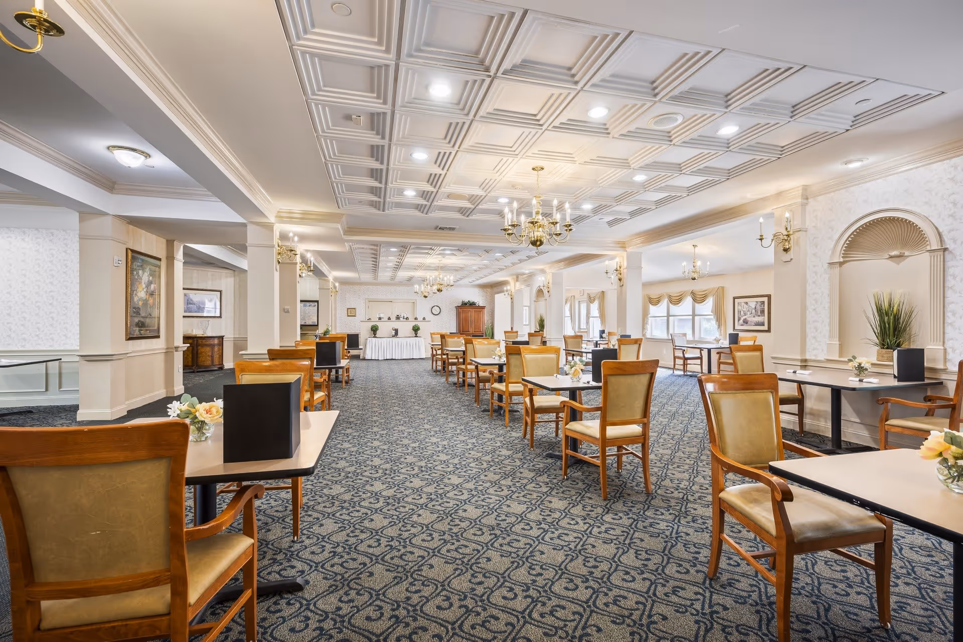 Spacious dining room with multiple tables and chairs arranged neatly, featuring patterned carpet, decorative ceiling panels with chandeliers, wall sconces, and large windows with curtains allowing natural light to fill the room.