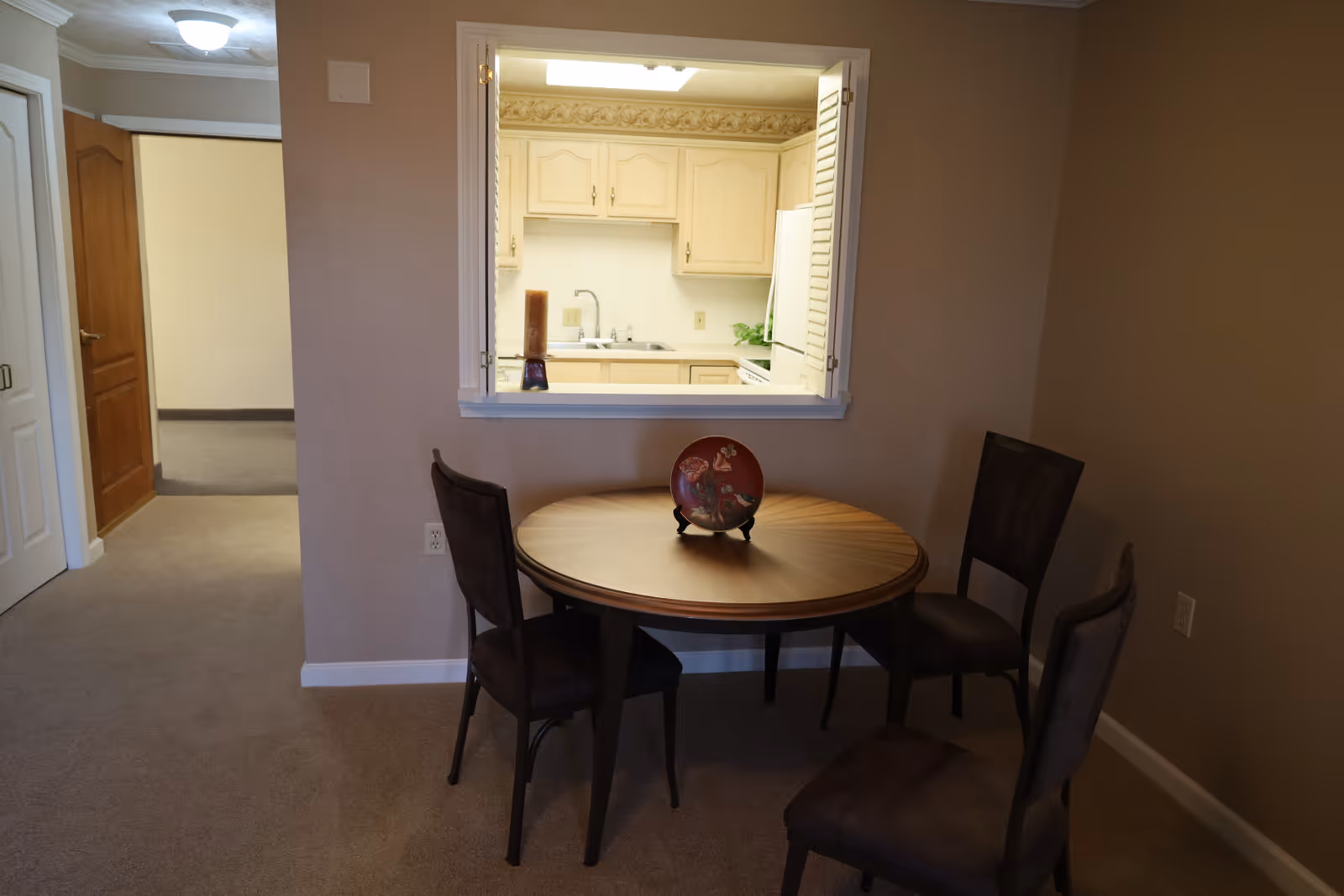 A small dining area with a round wooden table and three dark chairs. On the table is a decorative plate on a stand. The dining area is adjacent to a kitchen visible through a pass-through window, showing cream-colored cabinets and a white refrigerator. The walls are beige and the floor is carpeted.