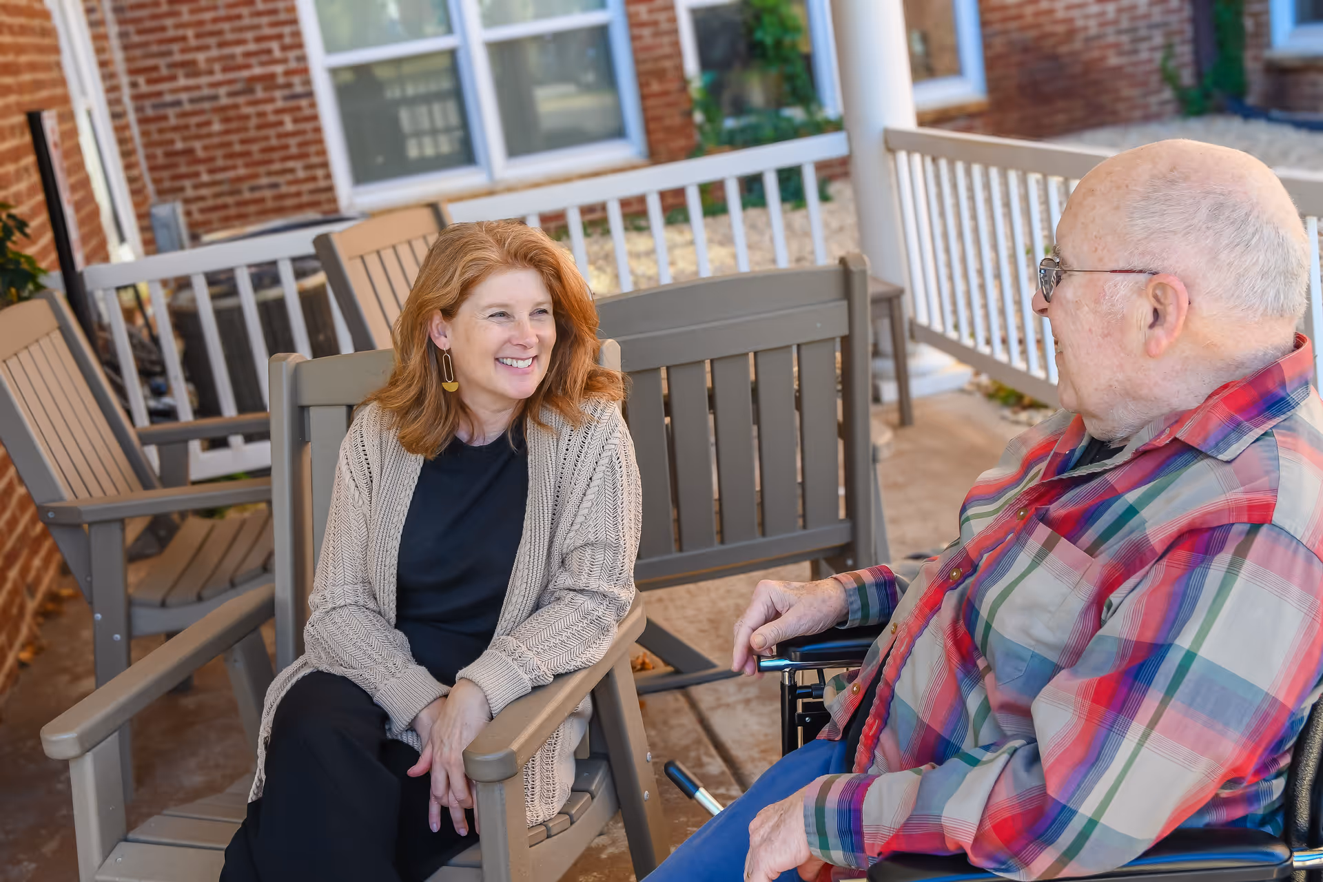 A woman with red hair wearing a beige cardigan and black pants sits on a wooden chair outdoors, smiling and talking to an elderly man in a wheelchair who is wearing glasses and a colorful plaid shirt. They are on a patio area with more wooden chairs and a brick building in the background.