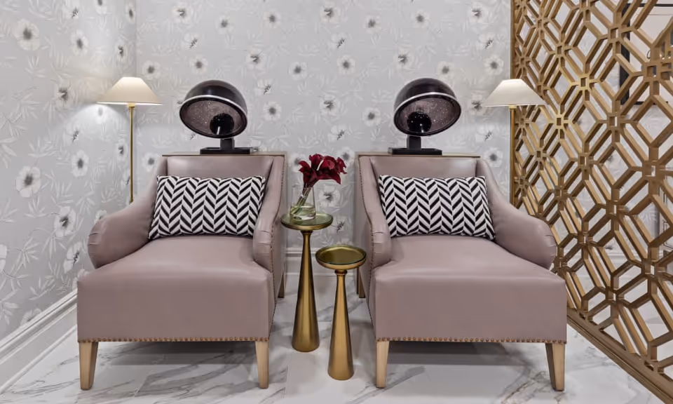Two mauve armchairs with black and white patterned cushions are placed side by side in a room with floral wallpaper. Between the chairs are two gold side tables, one holding a vase with red flowers. Behind each chair is a black hair dryer hood, and on either side of the chairs are tall floor lamps with white shades. A decorative wooden lattice screen is visible on the right side of the image.