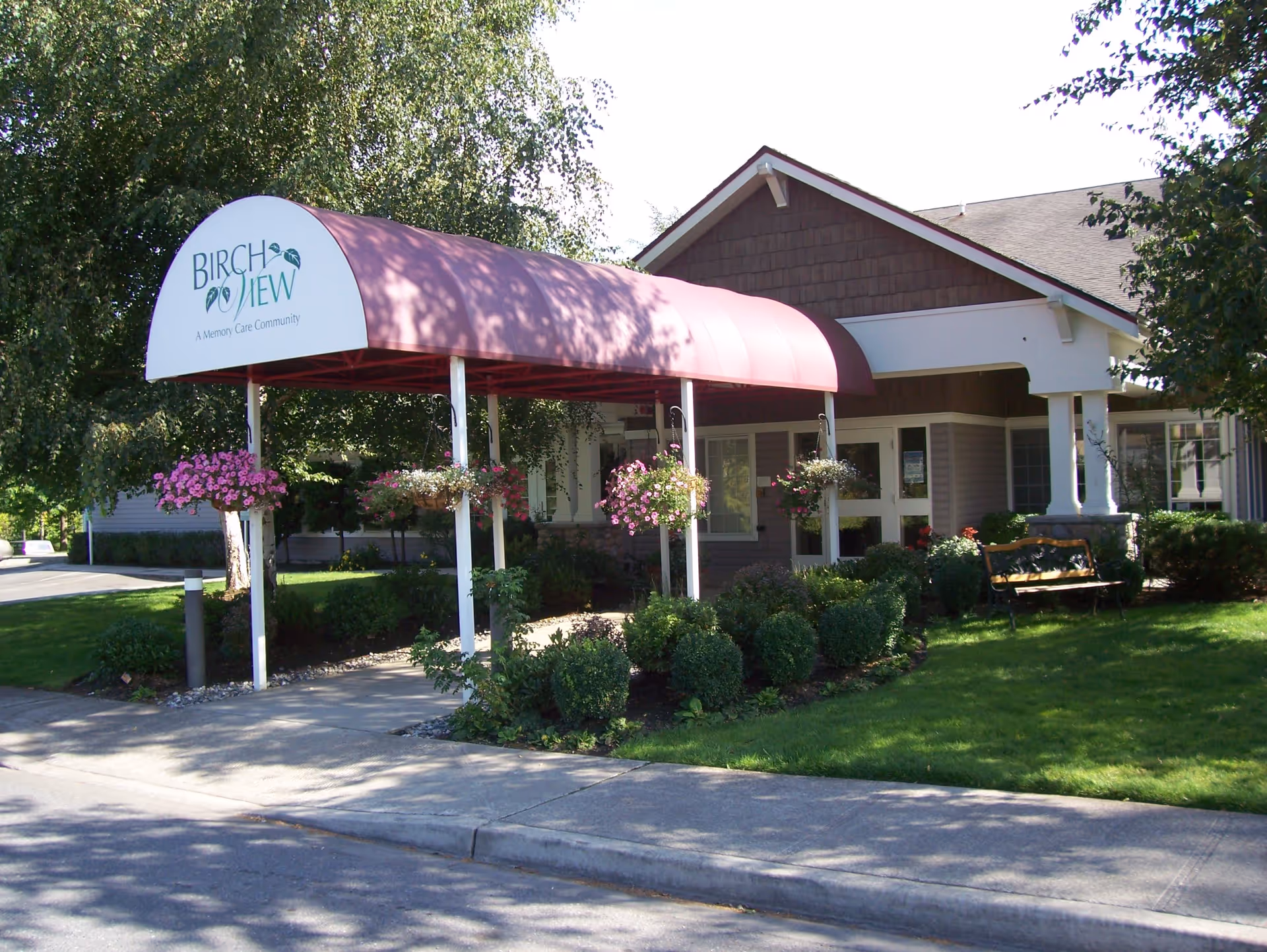 Exterior view of Birchview Memory Care facility entrance with a covered walkway featuring a maroon canopy and hanging flower baskets. The building has a brown roof, white pillars, and is surrounded by green shrubs and a lawn with a bench.