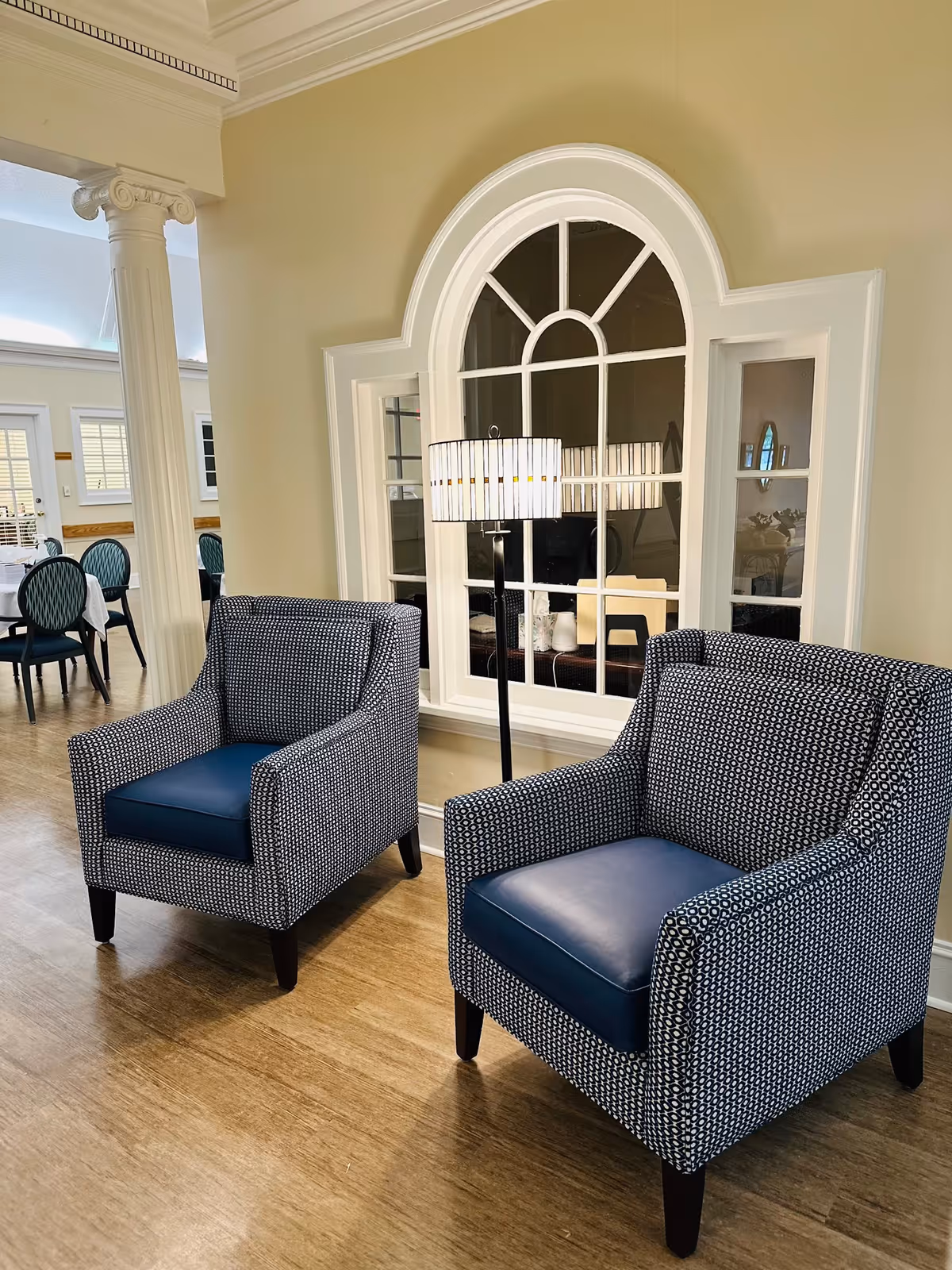 Two patterned armchairs with dark blue cushions are placed on a wooden floor in front of a large decorative window with an arched top. A tall floor lamp with a white shade stands between the chairs. In the background, there is a dining area with tables and chairs, and white columns supporting the ceiling.