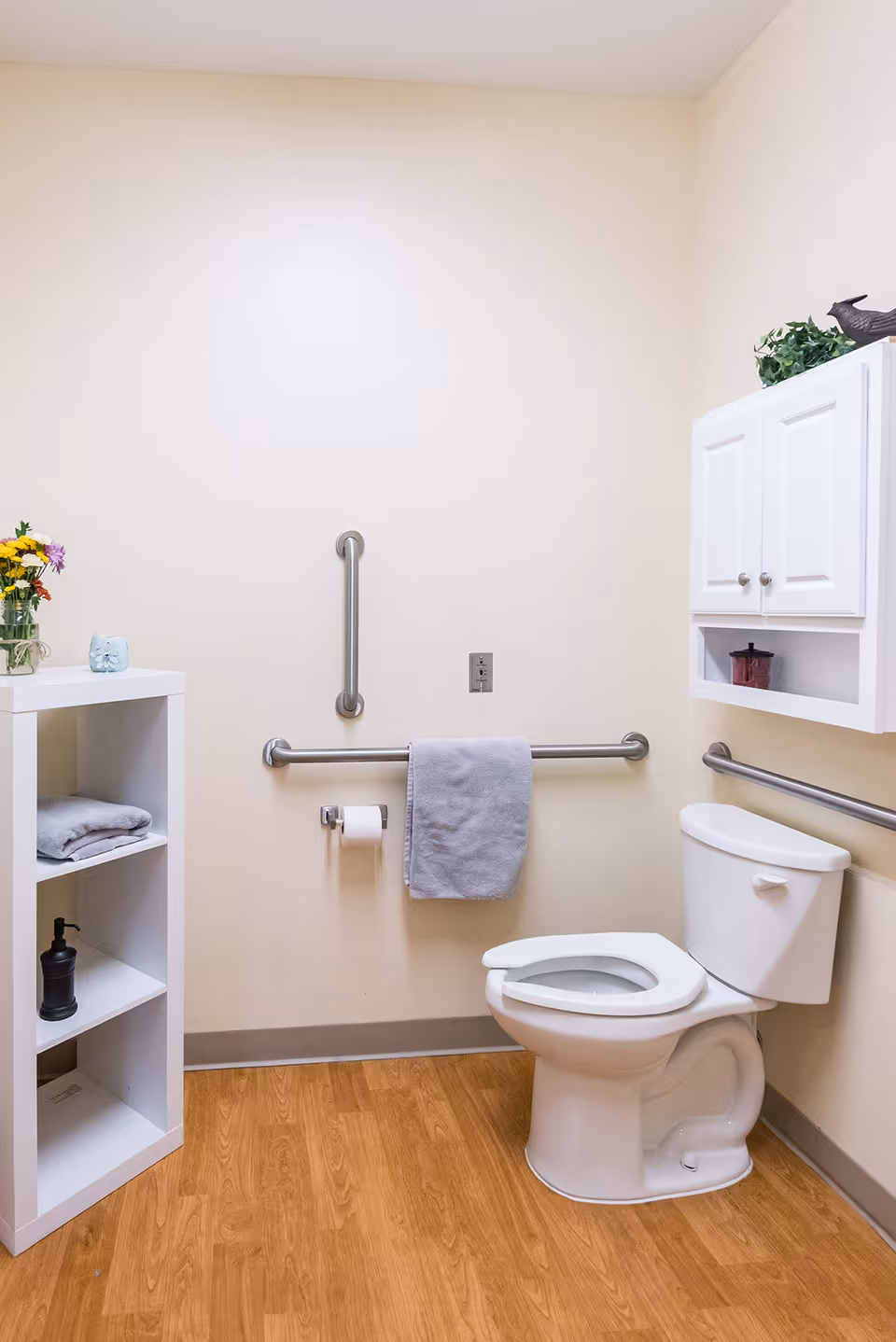 Accessible bathroom with a toilet, multiple grab bars, a towel on a rail, a wall cabinet and shelving on wood-look flooring.