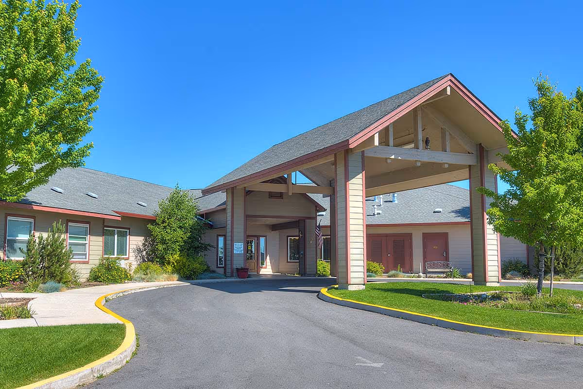 Exterior view of Eagle Lake Village senior living facility showing a covered entrance with a peaked roof, surrounded by green trees and landscaping under a clear blue sky.