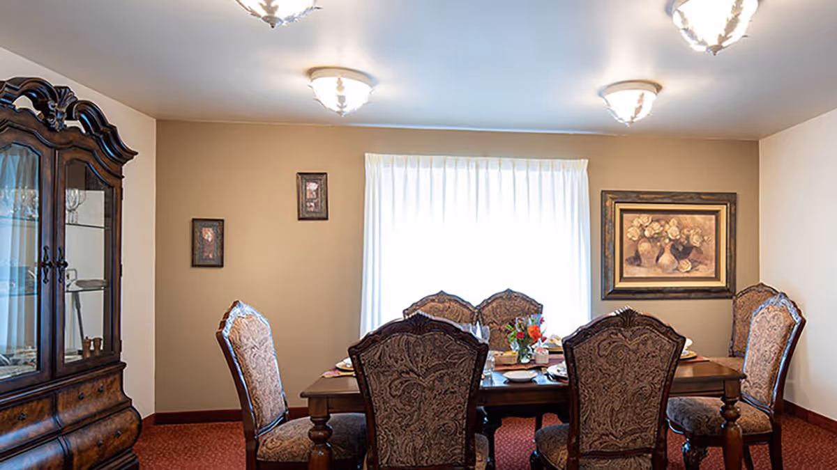 A dining room with a wooden dining table set for six people with ornate upholstered chairs. There is a wooden china cabinet on the left side and framed artwork on the walls. A window with white curtains is centered on the back wall, and three ceiling light fixtures illuminate the room.