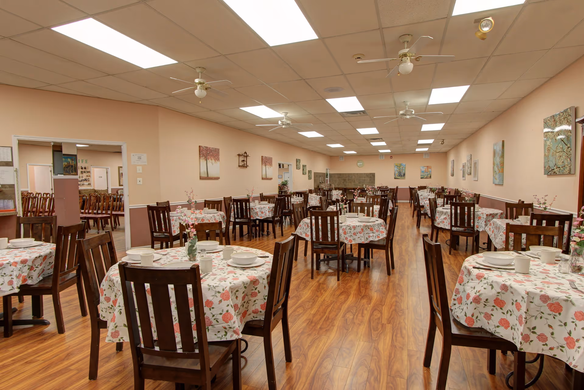 A spacious dining room with multiple round tables covered in floral tablecloths. Each table is set with white dishes, cups, and a small vase with pink flowers. The room has wooden flooring, beige walls adorned with paintings, and ceiling fans with lights. There is an open doorway leading to another room with more chairs.