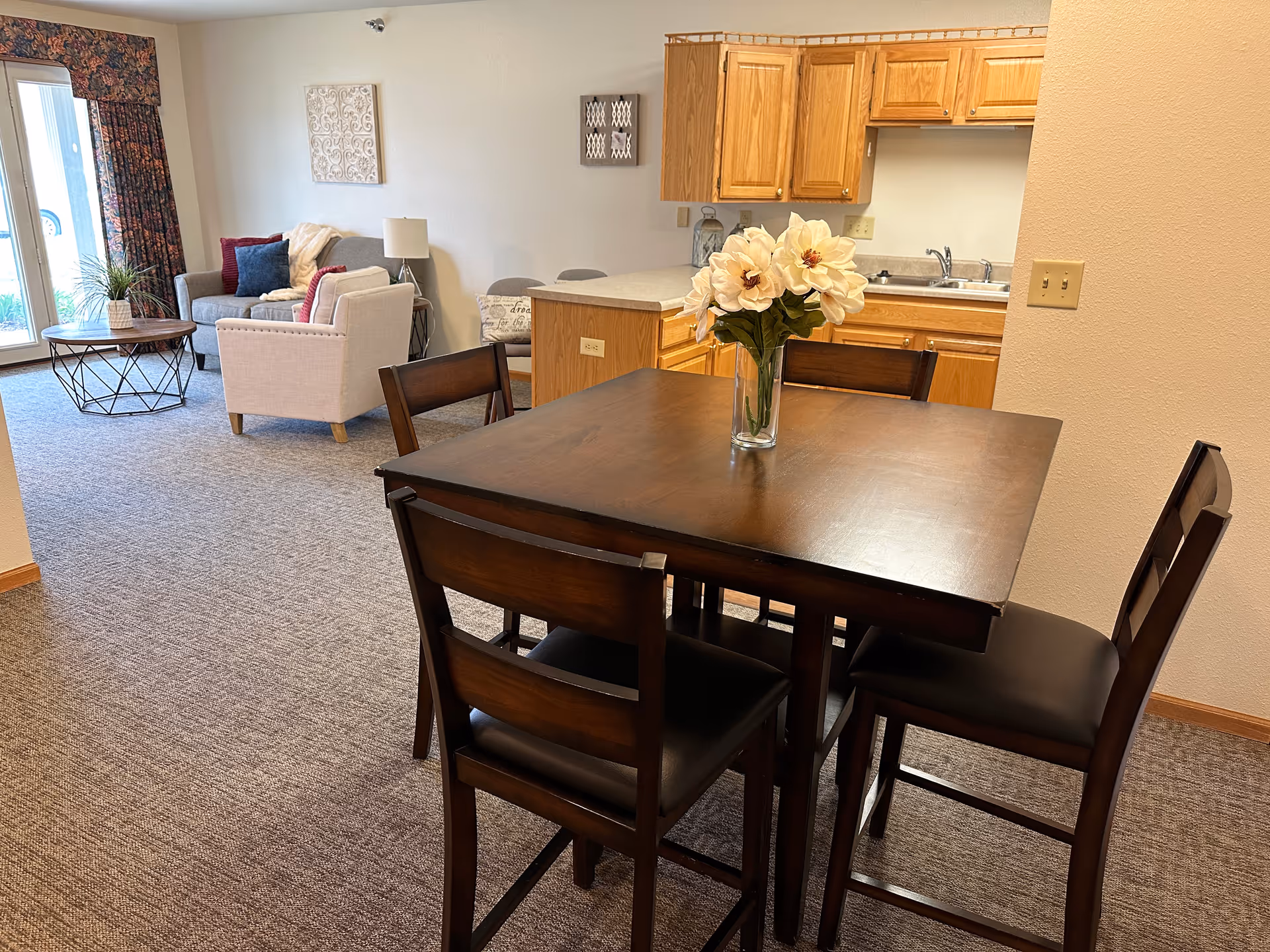 A cozy living and dining area in an assisted living facility. In the foreground, there is a dark wooden dining table with four matching chairs and a vase with white flowers on top. In the background, there is a small kitchen with wooden cabinets and a countertop. To the left, a living room area features a gray sofa, a beige armchair, a round coffee table, a floor lamp, and wall decorations. Large windows with floral curtains allow natural light into the room.