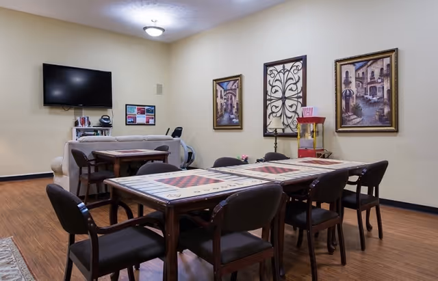 Communal activity room with game tables and chairs, a sofa and wall-mounted TV, framed artwork, and a popcorn machine.