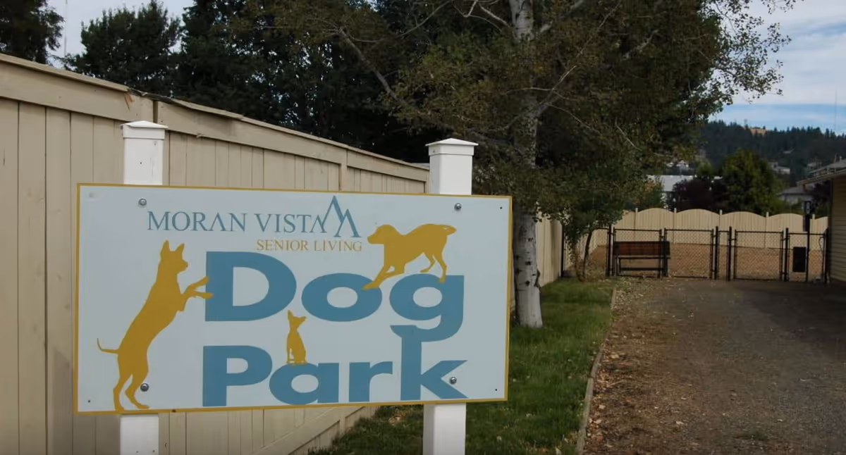 Outdoor dog park area at a senior living facility with a sign that reads 'Moran Vista Senior Living Dog Park' featuring silhouettes of dogs. The park is fenced with a bench inside and surrounded by trees and a beige fence.