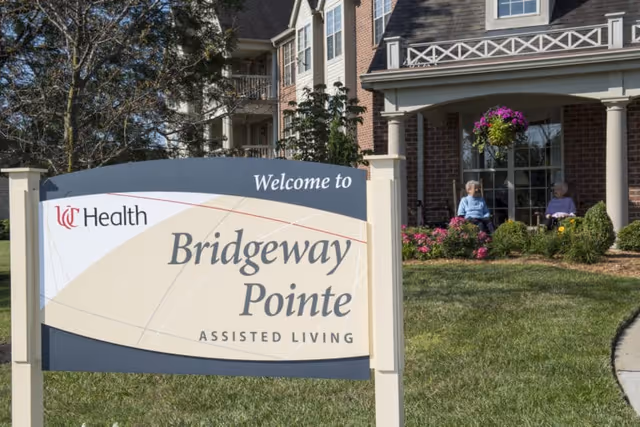 Outdoor view of the entrance area of Bridgeway Pointe assisted living facility with a welcome sign in the foreground and two elderly women sitting on chairs near the building surrounded by greenery and flowers.