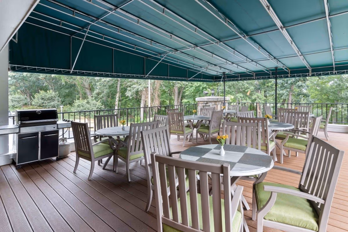 Covered outdoor patio area with multiple round tables and wooden chairs with green cushions. Each table has a small vase with yellow flowers. There is a black grill on the left side and a wooded area visible beyond the railing.
