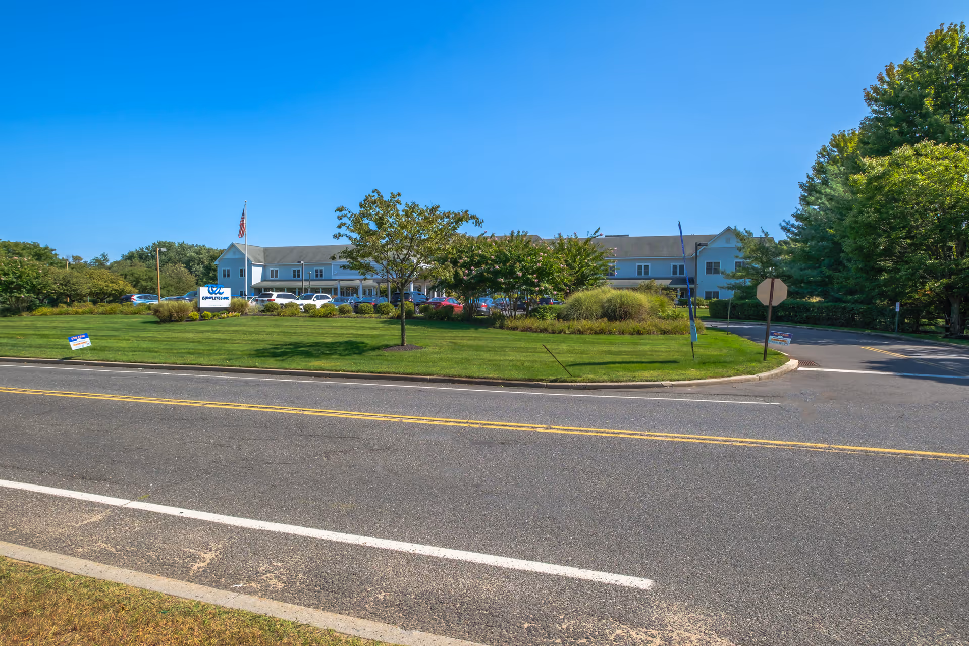 Front exterior of the Complete Care at Holmdel building with a manicured lawn, flagpole and parking area visible behind a roadway.