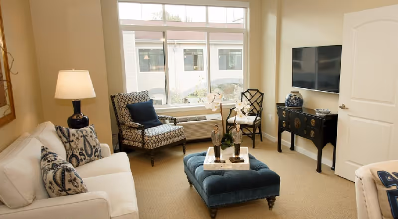 A cozy living room with a white sofa adorned with patterned pillows, a patterned armchair with a navy pillow, a blue upholstered ottoman with decorative figurines on top, a black sideboard with a blue and white vase, and a wall-mounted flat-screen TV. The room has beige walls and carpet, a large window letting in natural light, and a table lamp on a side table next to the sofa.