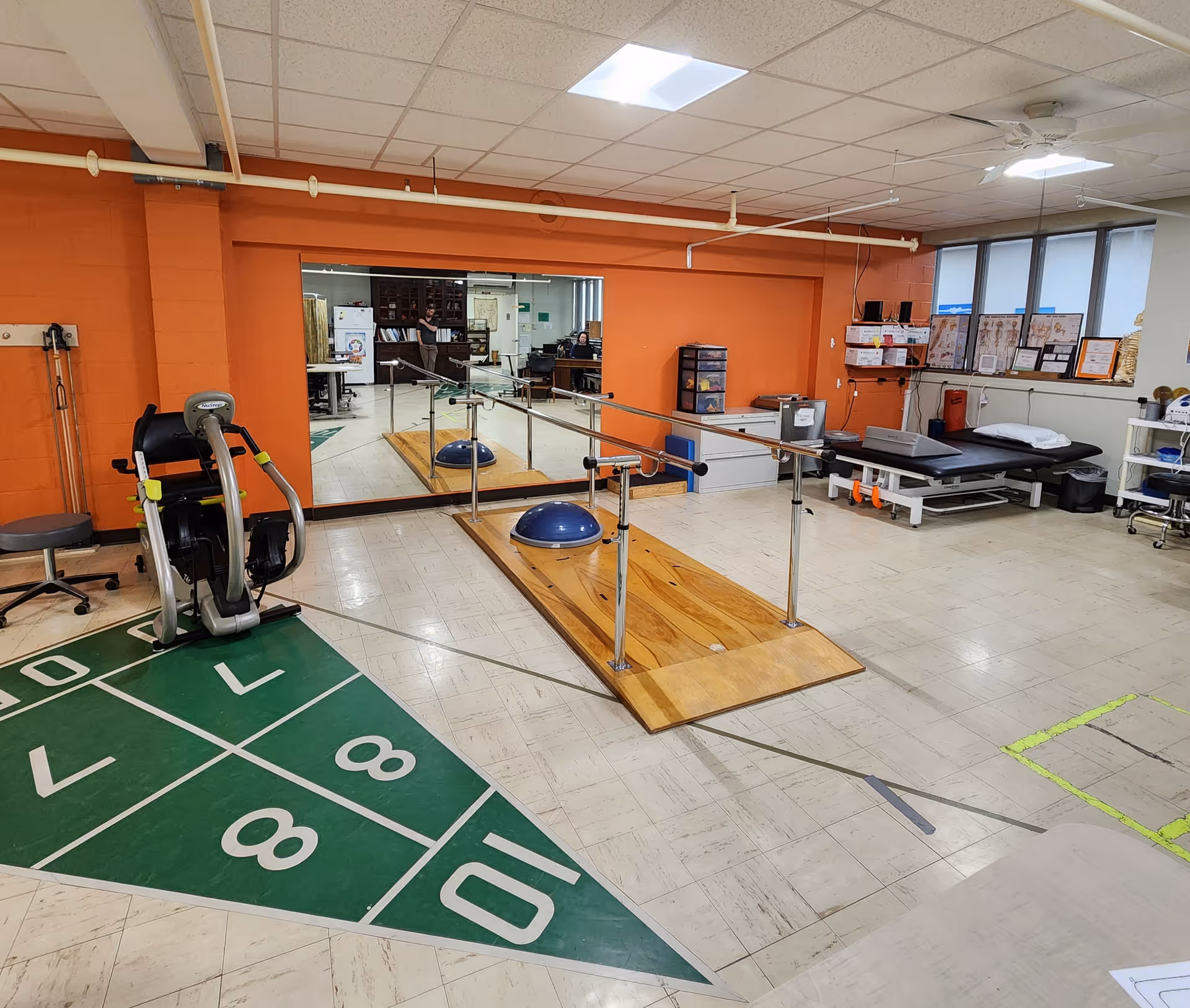 Physical therapy room with parallel bars for walking exercises, a green shuffleboard court painted on the floor, exercise equipment, a treatment table with a pillow, anatomical charts on the wall, and a large mirror reflecting the room.