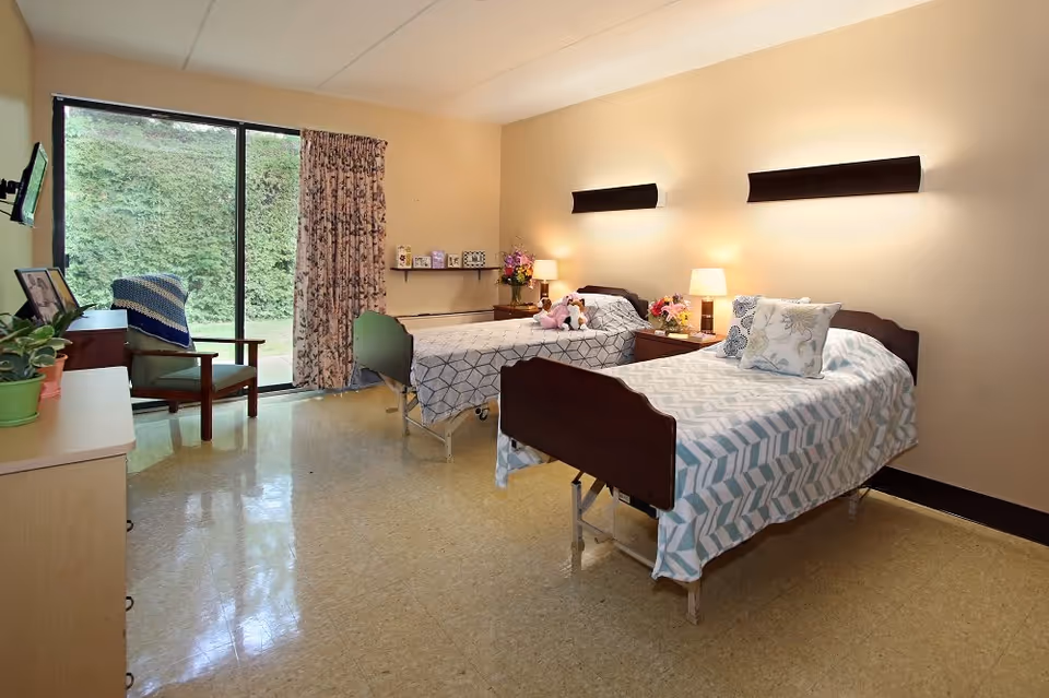 A senior living facility bedroom with two single beds, each with patterned bedspreads and pillows. There are nightstands with lamps and flowers between the beds. A chair with a blanket is near a sliding glass door with floral curtains, leading to an outdoor area with greenery. A dresser with plants and framed photos is on the left side of the room.
