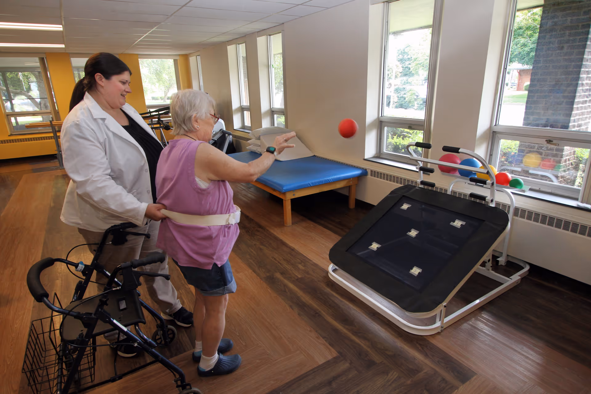 An elderly woman with a walker is playing a ball toss game with the assistance of a caregiver in a rehabilitation room with large windows and wooden flooring.