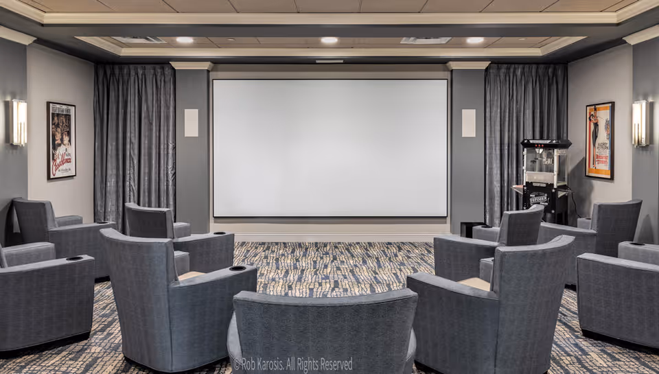 A small theater room with multiple gray upholstered armchairs arranged in rows facing a large blank projection screen. The room has patterned carpet, gray walls with framed movie posters, and a popcorn machine in the corner.