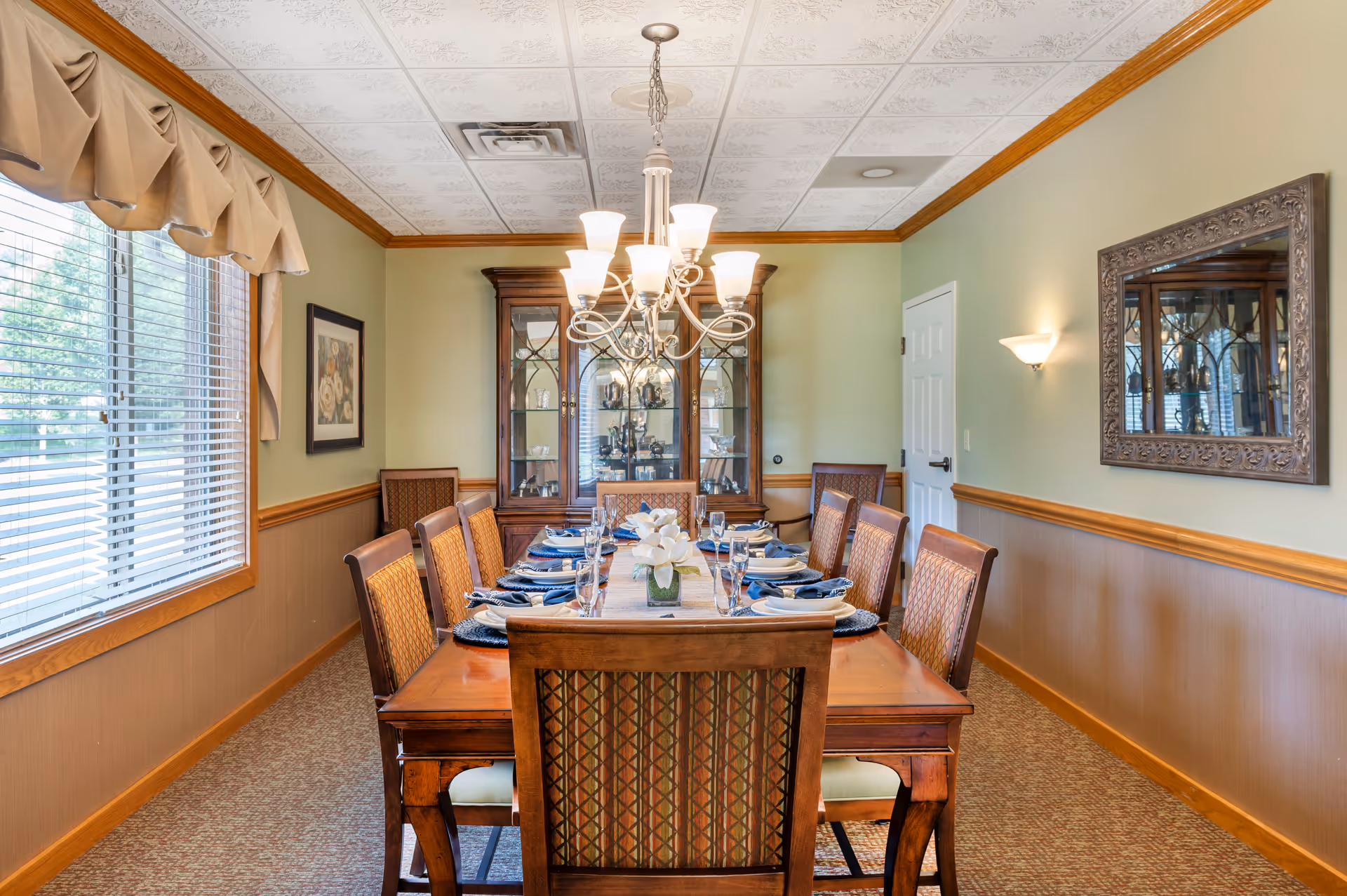 A formal dining room with a long wooden table set for eight people. The table is arranged with blue napkins, plates, glasses, and a white floral centerpiece. The room features a large window with blinds and a valance on the left, a wooden china cabinet with glass doors at the far end, a decorative mirror on the right wall, and a chandelier hanging above the table. The walls are painted light green with wooden wainscoting and crown molding.