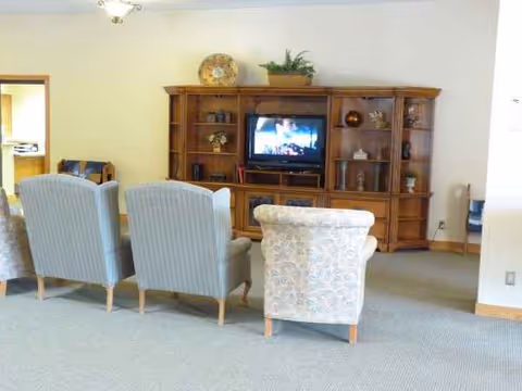A senior living facility lounge with three light chairs facing a wooden entertainment center and TV