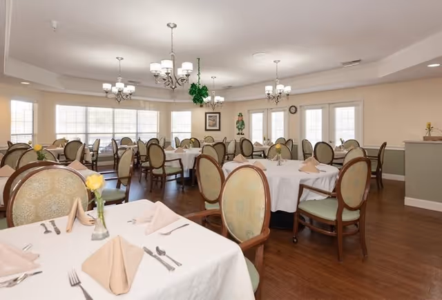 A bright dining room with multiple round tables covered with white tablecloths, each set with beige folded napkins, silverware, and small vases holding yellow flowers. The room features wooden chairs with cushioned seats and backs, large windows allowing natural light, and elegant chandeliers hanging from the ceiling.
