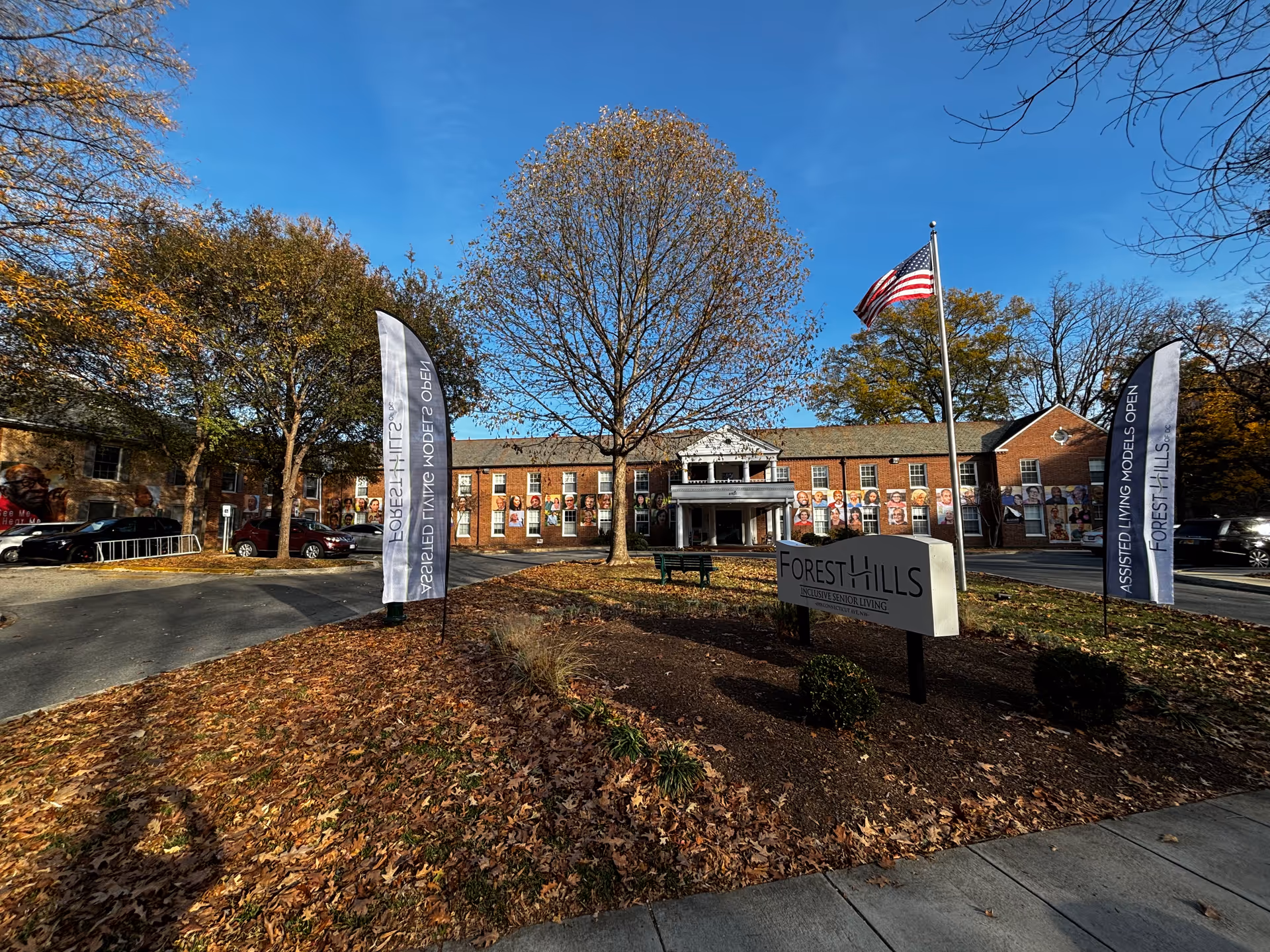 Exterior view of Forest Hills inclusive senior living facility with a brick building, an American flag on a pole, two tall banners advertising assisted living models open, and a sign with the facility name. Trees with autumn leaves surround the area under a clear blue sky.