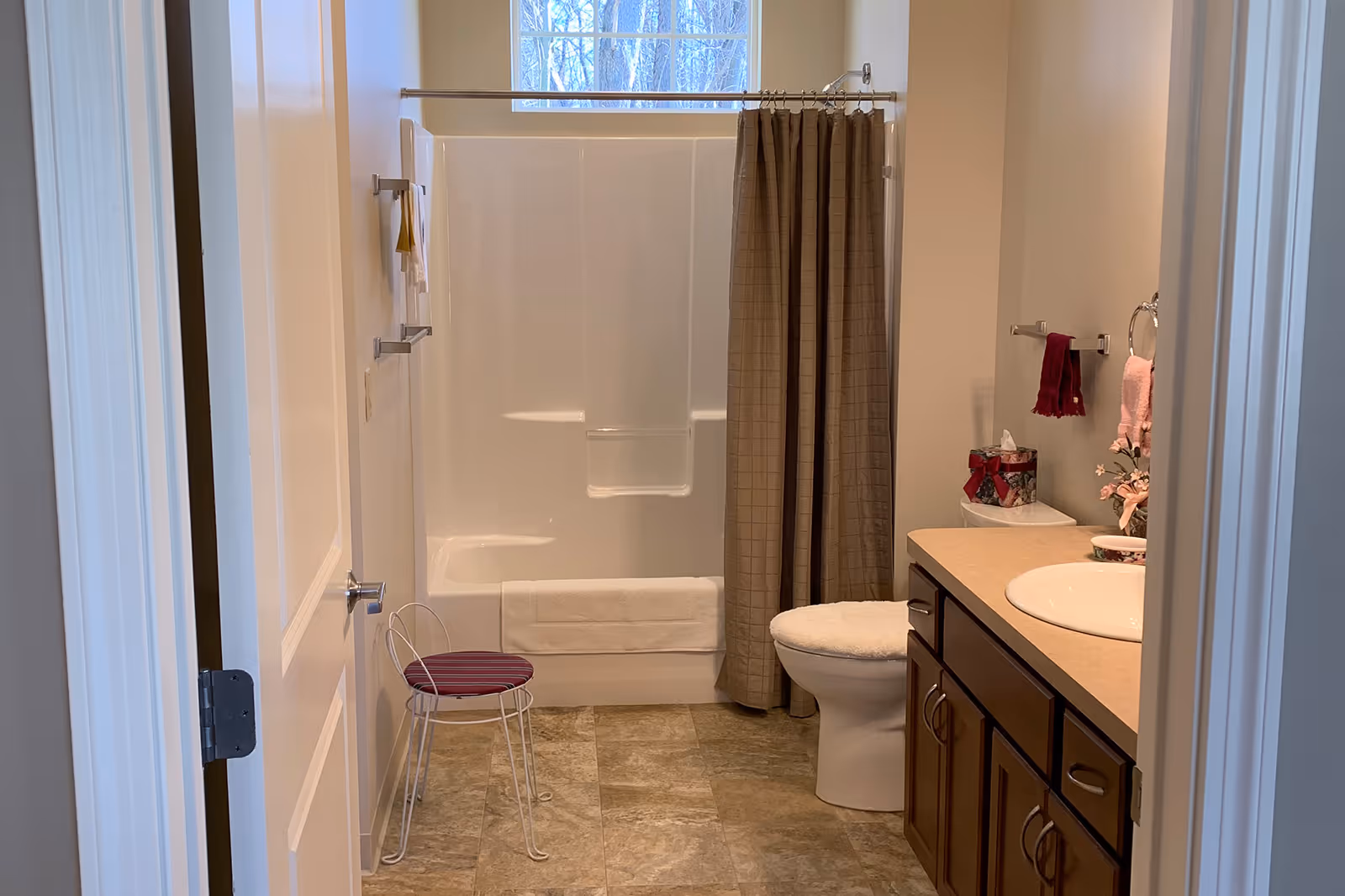 A clean bathroom with a white bathtub and shower combination with a beige shower curtain, a white toilet with a cushioned seat cover, a wooden vanity with a sink, and a small white metal stool with a red cushion. There are towels hanging on the wall and a window above the bathtub letting in natural light.