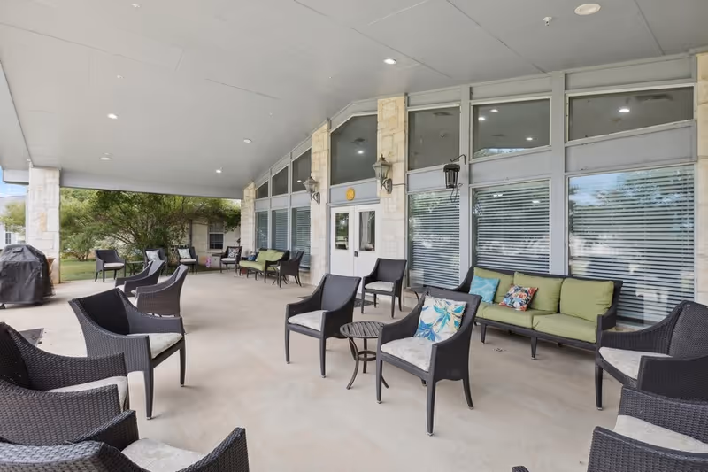 Covered outdoor patio area with multiple black wicker chairs and green cushioned sofas arranged around small tables. The patio is adjacent to a building with large windows and double doors, surrounded by stone pillars and outdoor lantern-style lights. Trees and greenery are visible in the background.