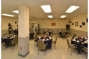 Interior view of a dining room with several round tables covered with dark tablecloths, each set with plates, cups, and napkins. The room has beige walls with a two-tone paint design, fluorescent ceiling lights, and some framed pictures and plants on the walls.