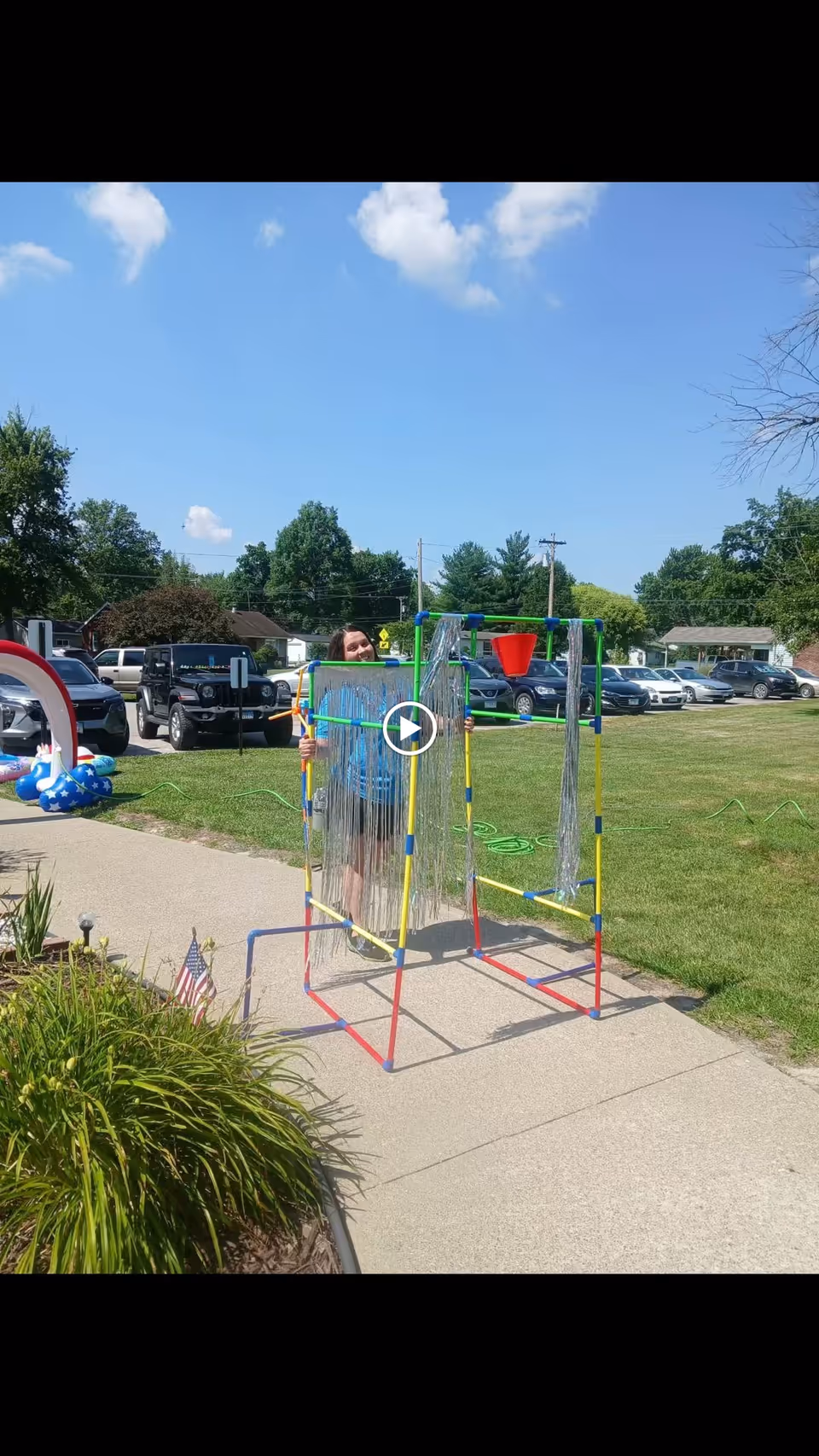 A person standing outdoors on a sidewalk next to a colorful frame structure decorated with silver streamers and a red bucket on top. There are parked cars and green grass in the background under a blue sky with some clouds.
