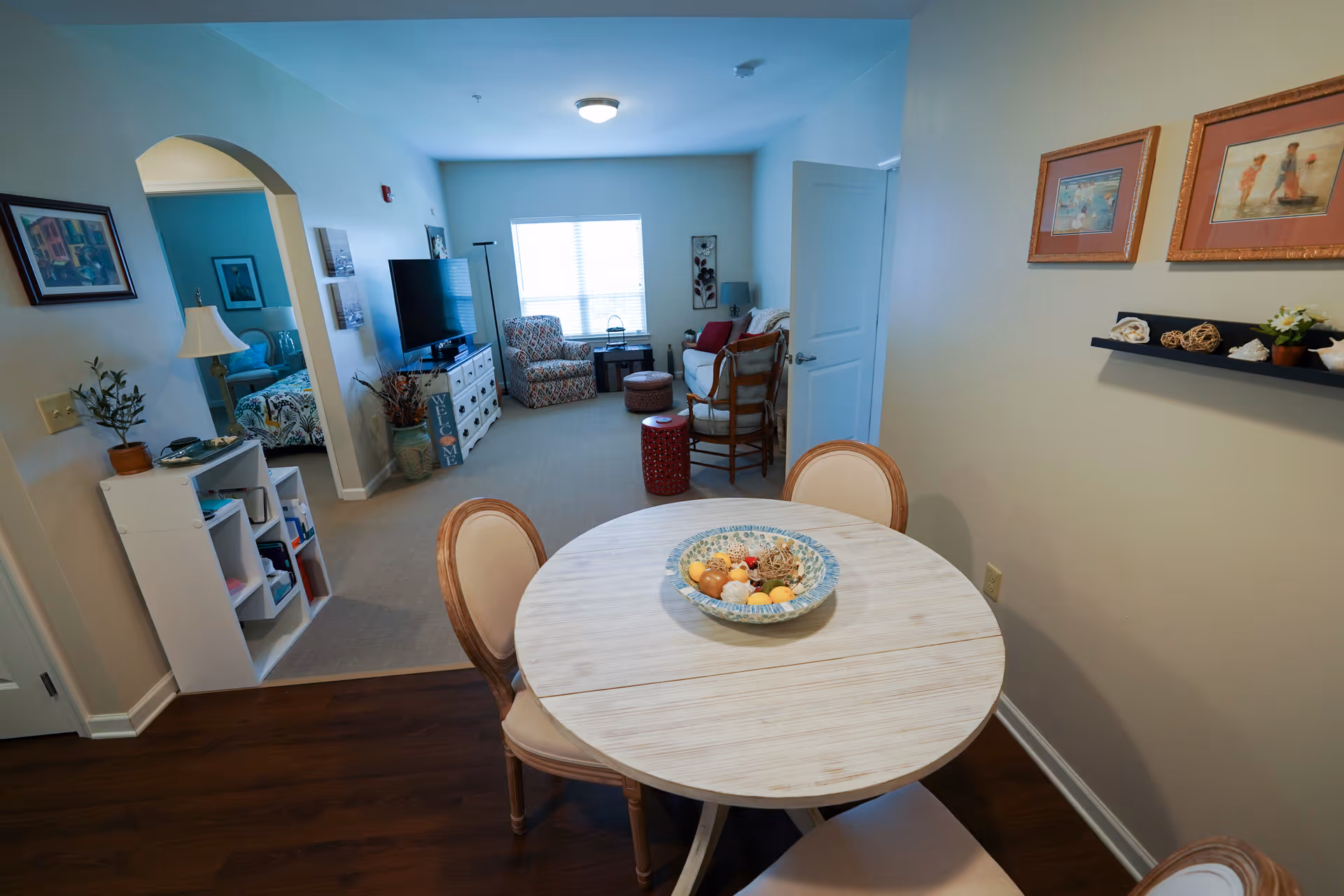 Bright combined dining and living area with a round wooden table in the foreground and a TV and seating by a window in the background.