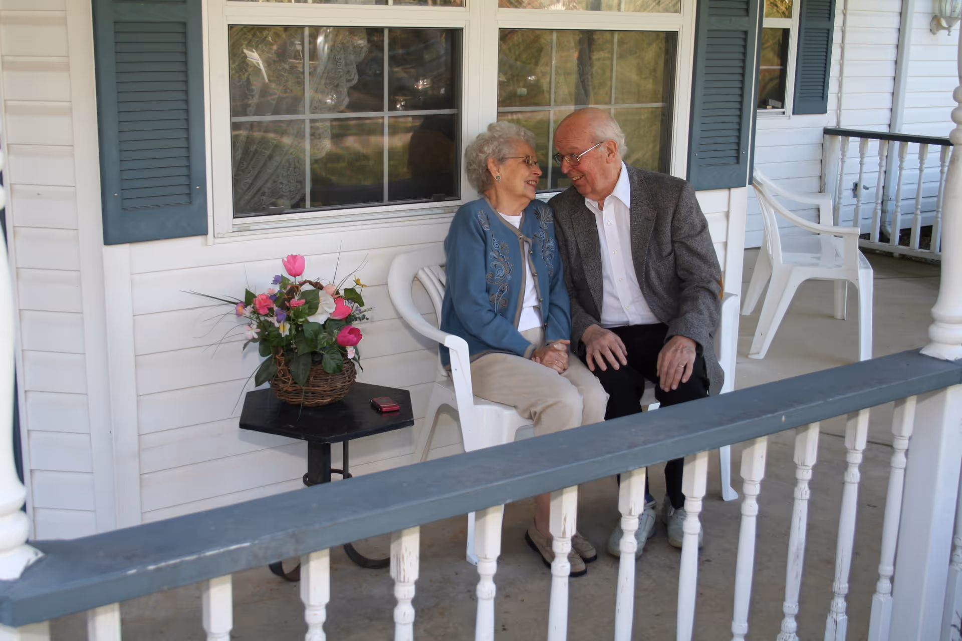 An elderly couple sitting closely together on white plastic chairs on a porch, smiling and holding hands. There is a small black table next to them with a basket of colorful flowers and a small red object. The porch has white railings and a window with blue shutters behind them.