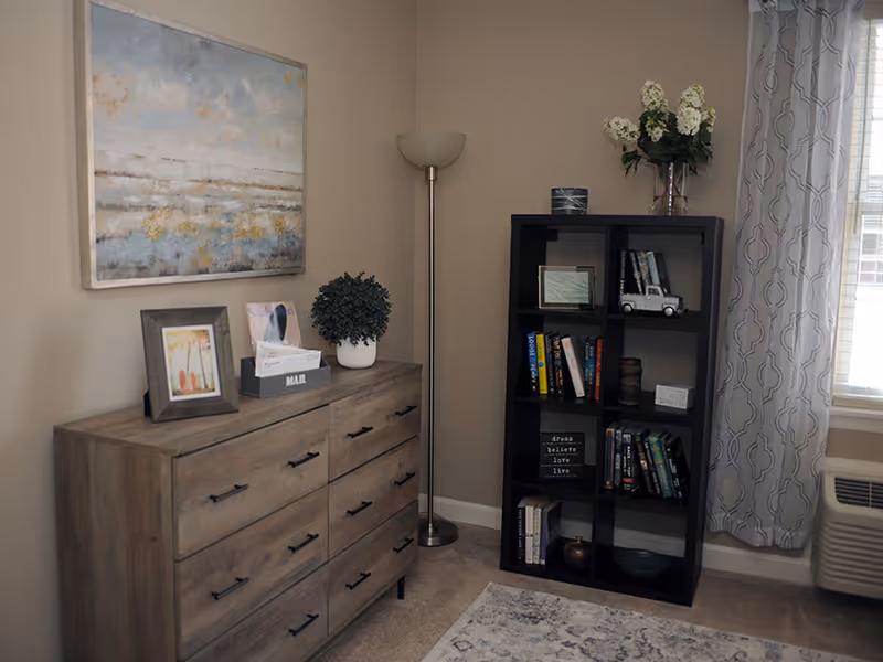 A cozy corner of a room featuring a wooden dresser with framed pictures, a mail organizer, and a potted plant on top. Next to it is a tall black bookshelf filled with books, decorative items, and a vase with white flowers on top. A floor lamp stands between the dresser and bookshelf. A window with patterned curtains and a heating/cooling unit is visible on the right side.