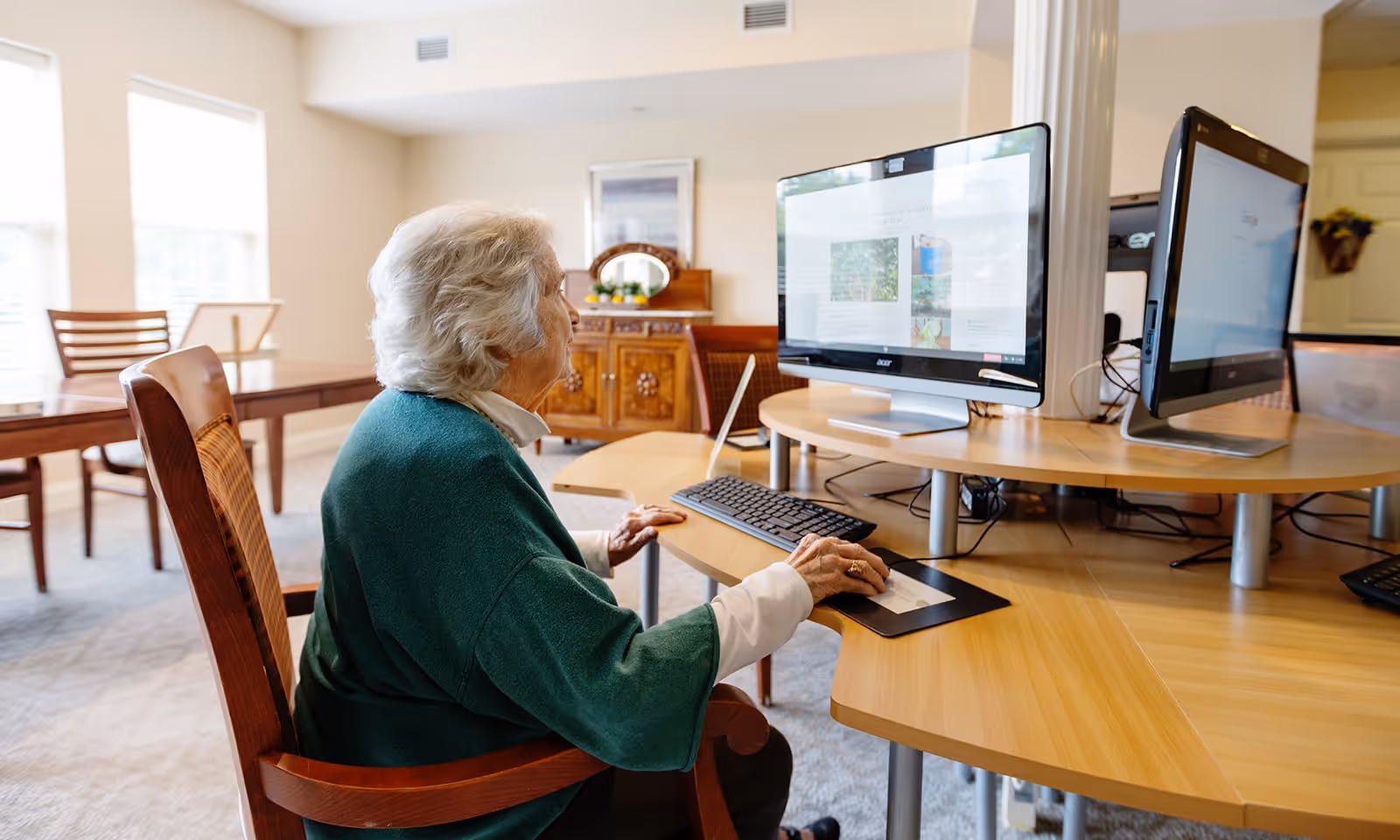 An elderly woman with gray hair wearing a green sweater is sitting at a wooden desk using a desktop computer in a well-lit room with large windows and wooden furniture.
