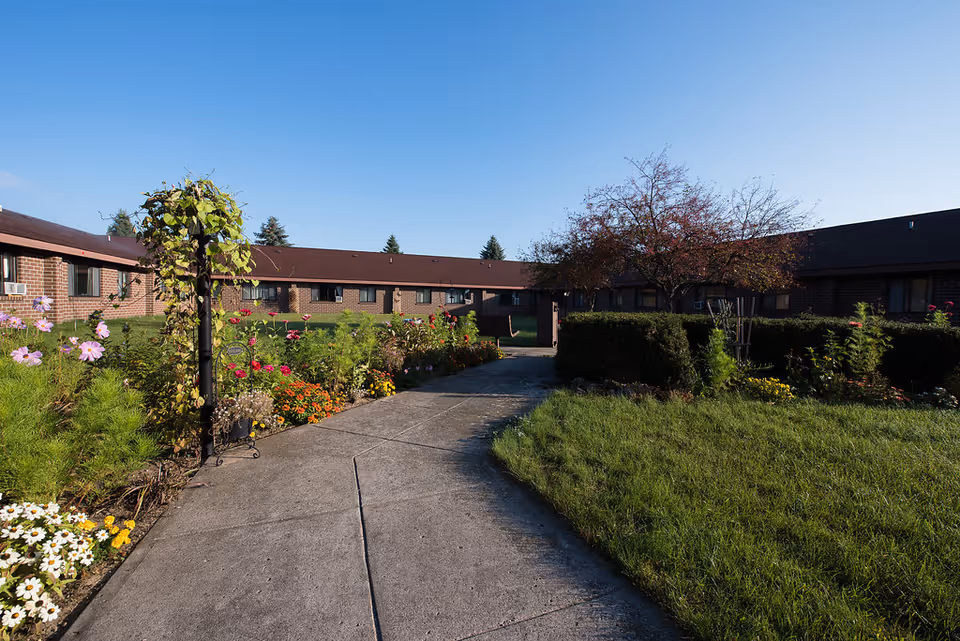 A paved courtyard walkway lined with flower beds leading toward a low brick single-story building under a clear blue sky.