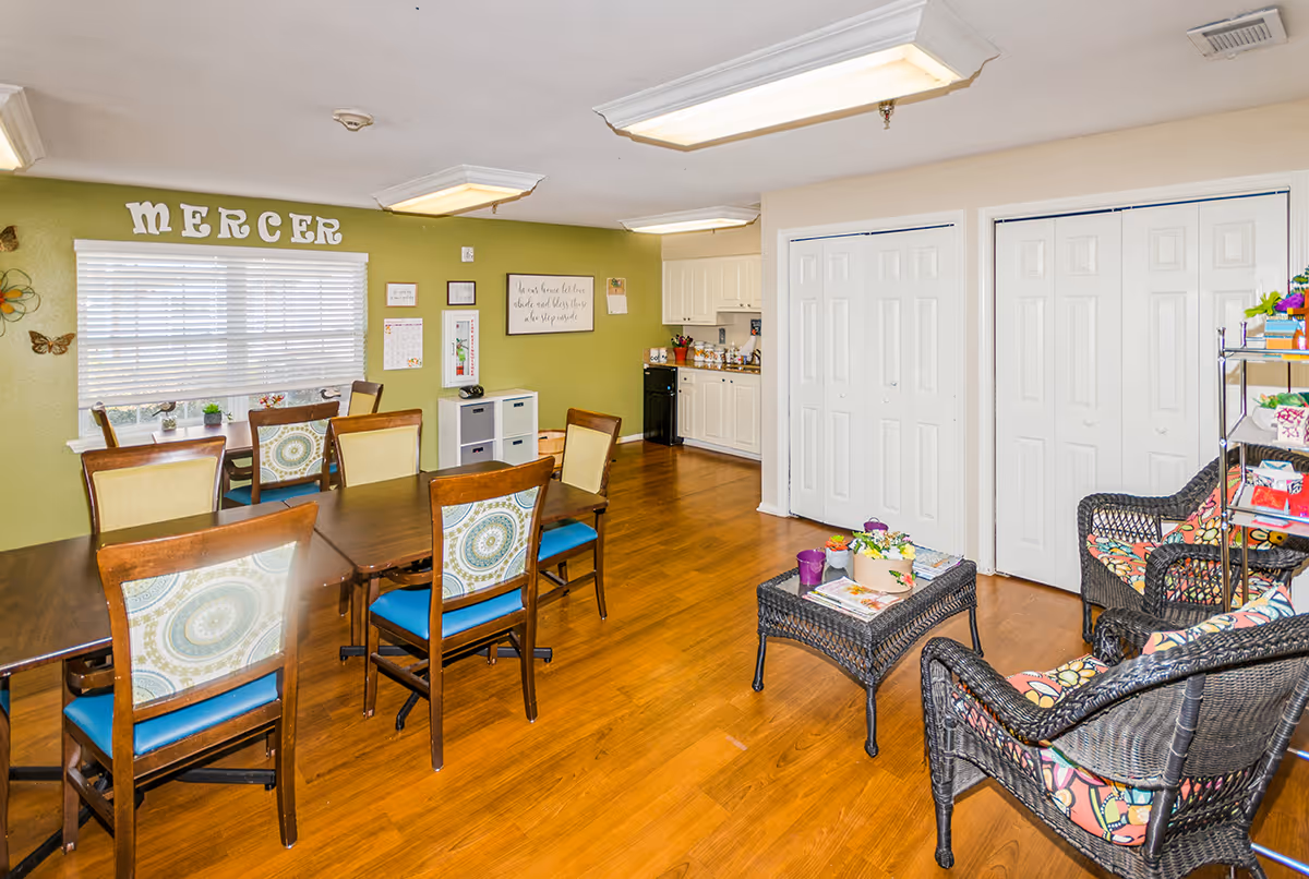 A bright and cozy common area in Mercer Terrace featuring a dining table with six chairs, a small kitchen area with white cabinets and a black dishwasher, and a seating area with two wicker chairs and a wicker coffee table with magazines and decorative items. The walls are painted green and white, with the word 'MERCER' displayed above a window with white blinds. The floor is wooden, and there are ceiling lights providing illumination.