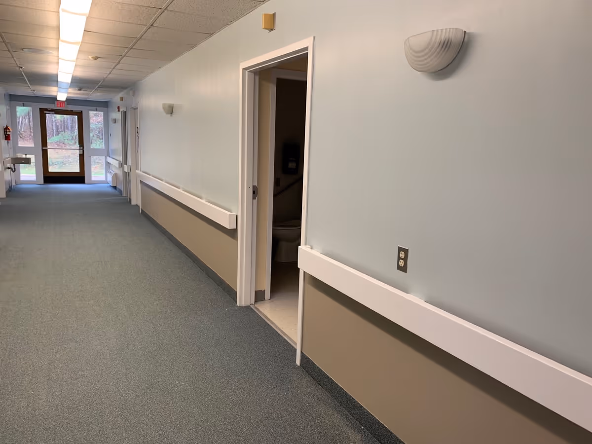 A long indoor hallway with light blue and beige walls, carpeted floor, and ceiling lights. On the right side, there is an open door revealing a bathroom with a visible toilet. At the end of the hallway, there is a glass door leading outside with trees visible through the glass.