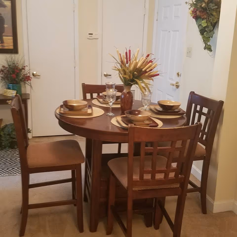 A round wooden dining table set for four with matching wooden chairs. Each place setting includes a bowl, plate, and glass. A vase with decorative dried flowers is in the center of the table. The room has beige walls and carpet, with two white doors and a small table with plants and decor in the background.