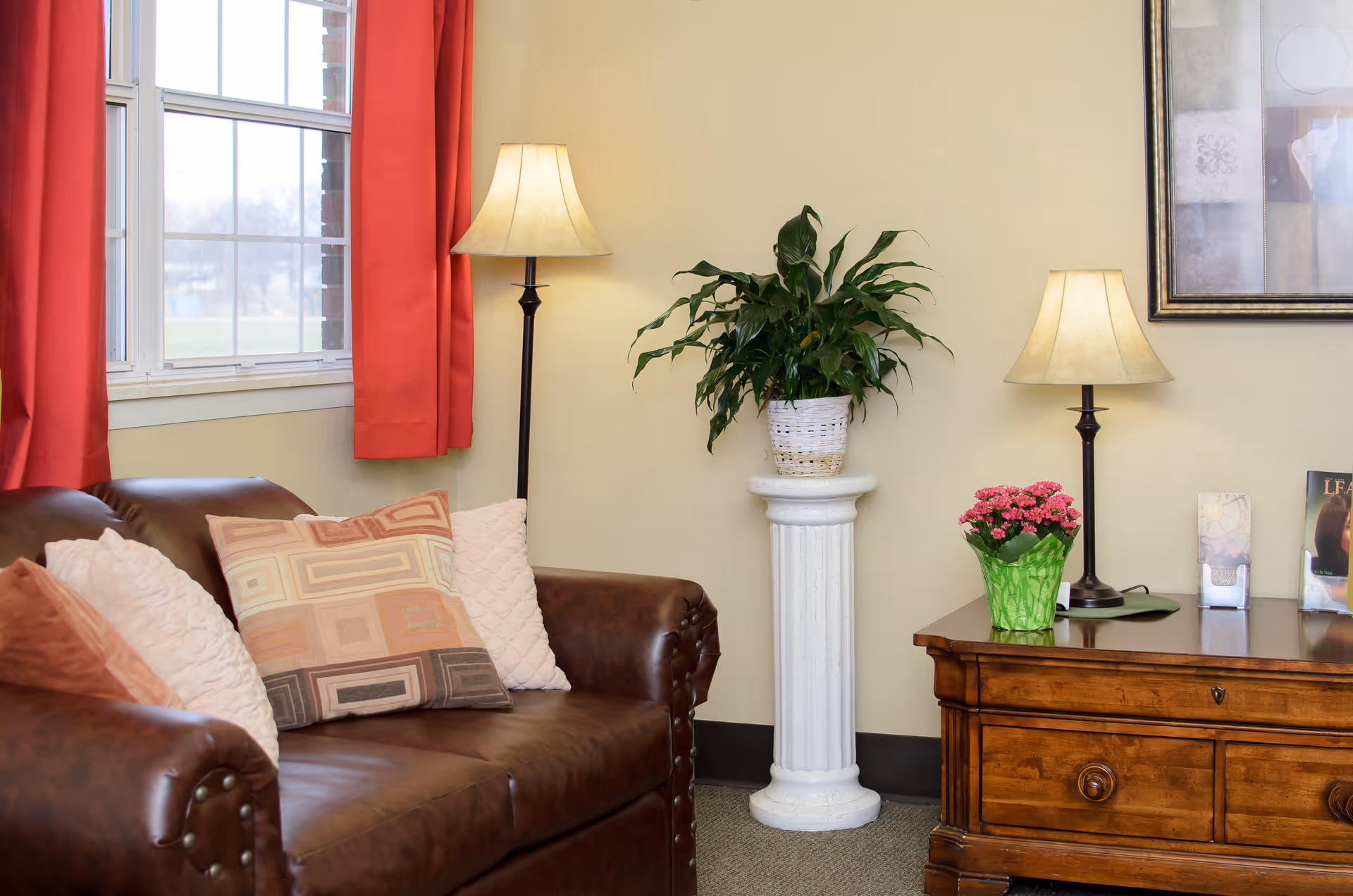 A cozy living room area with a brown leather sofa adorned with patterned and textured pillows. Next to the sofa is a tall floor lamp with a beige lampshade. A white pedestal holds a green leafy plant in a white basket. To the right, there is a wooden side table with a small table lamp, a pink flowering plant in a green pot, and some brochures or magazines. The walls are light yellow, and there is a window with red curtains letting in natural light.