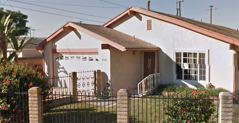 Exterior view of a single-story residential building with a white facade and pink trim. The building has a garage door, a front door with a small ramp, and a window with bars. There is a fenced front yard with green grass, bushes, and flowering plants.