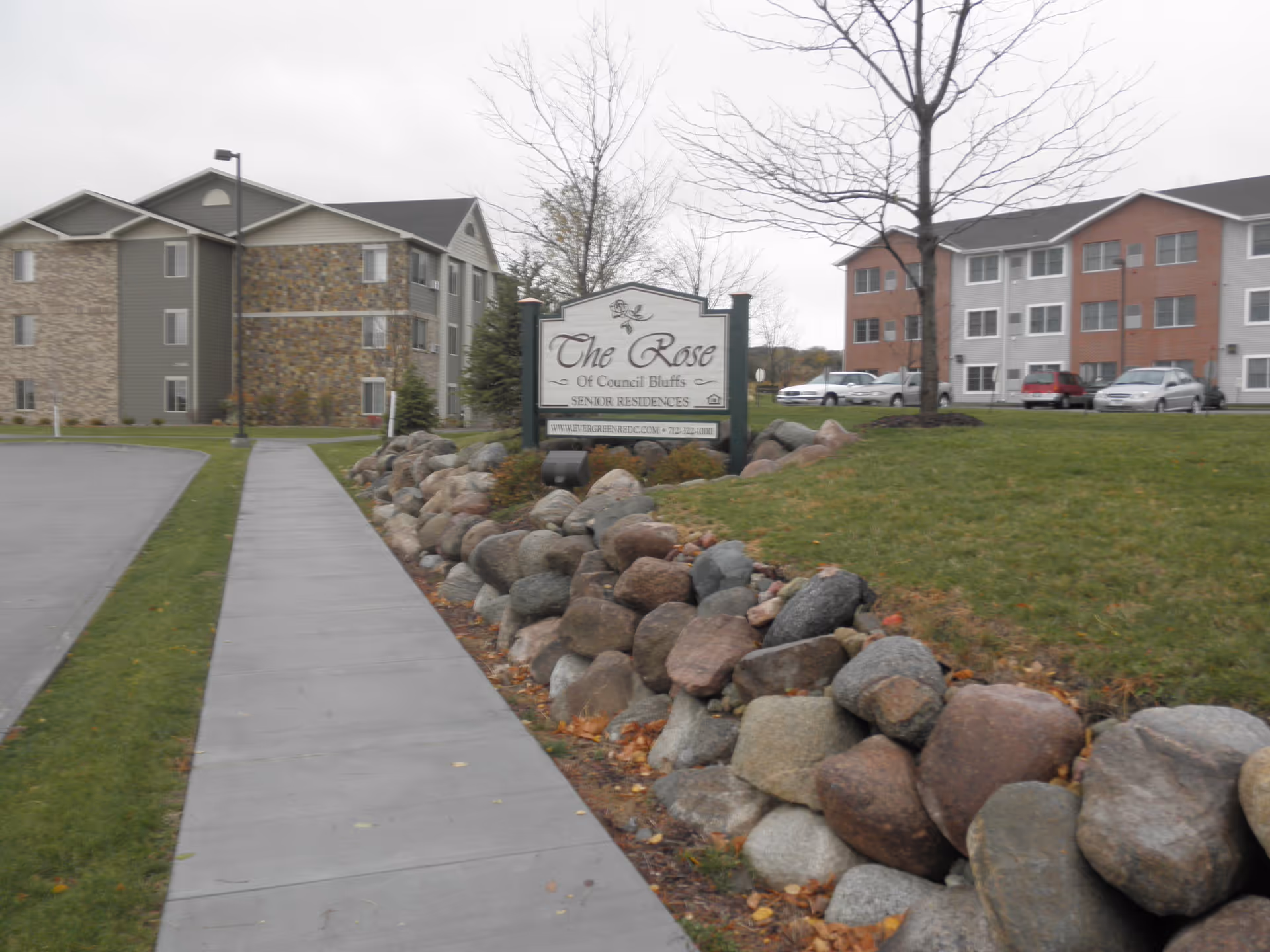 Outdoor view of The Rose of Council Bluffs senior residences showing a sidewalk bordered by a rock garden and grass, with multi-story residential buildings and parked cars in the background under an overcast sky.