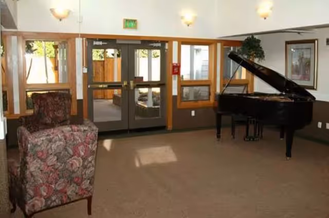 A common area with a floral upholstered armchair on the left and a black grand piano on the right. The room has beige carpet, white walls with brown trim, and three wall-mounted light fixtures. Double glass doors lead outside, with windows on either side allowing natural light in.