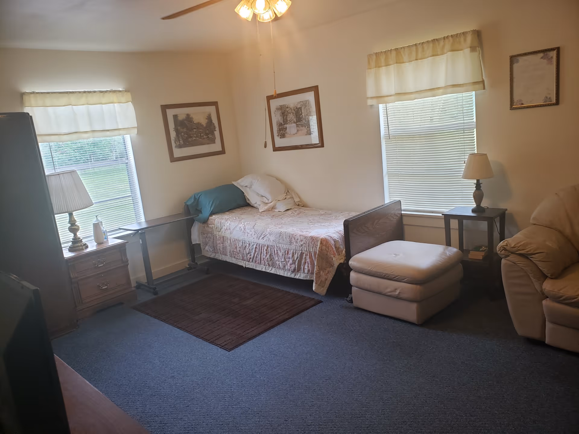 Bedroom in a senior living facility with bed, ottoman, lamps, and windows.