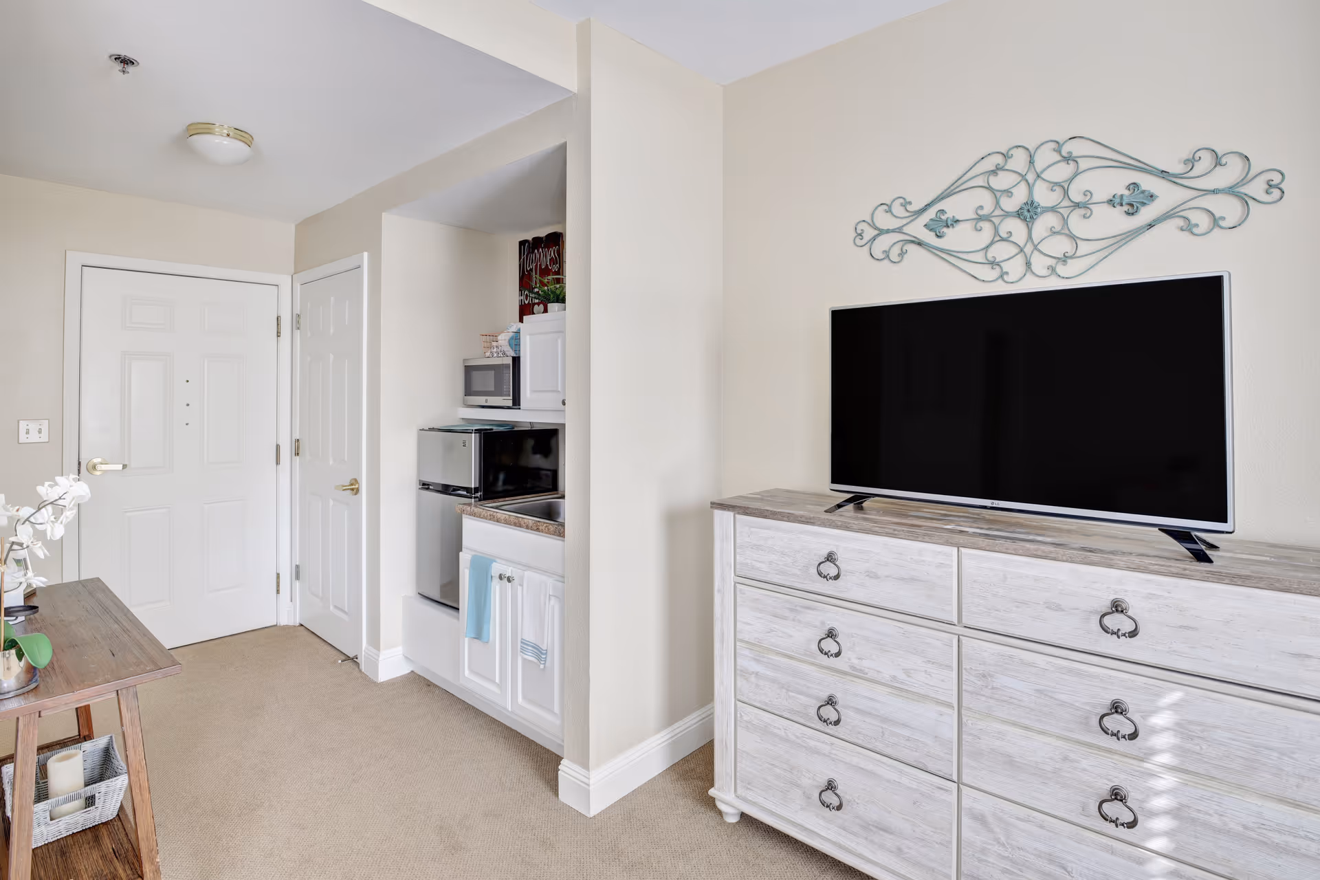 Interior view of a senior living facility room showing a white door, a small kitchenette with a microwave, mini fridge, and sink, a wooden dresser with a flat-screen TV on top, and a decorative metal wall art above the TV.