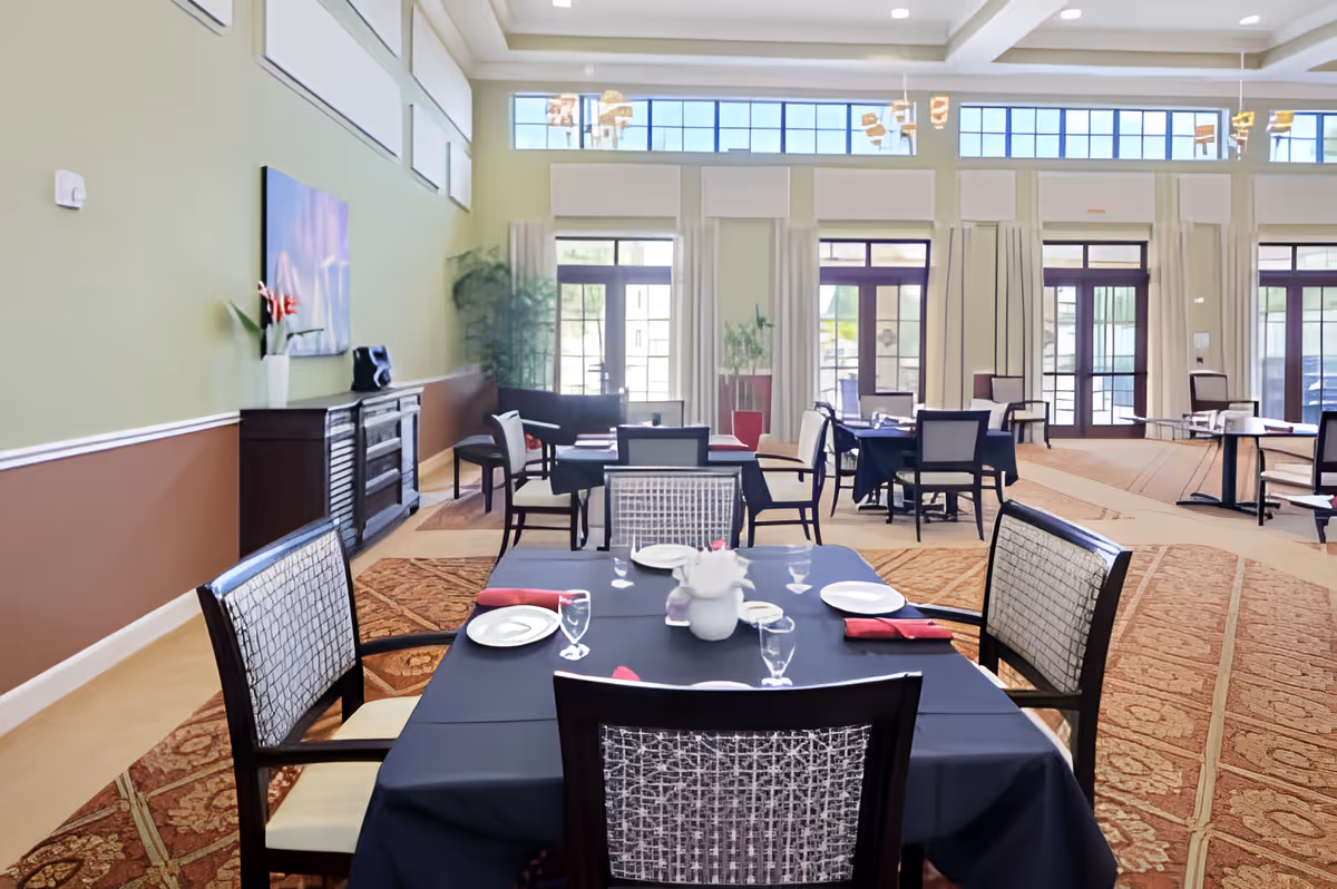 A spacious dining room with several tables covered in dark tablecloths, each set with plates, glasses, and red napkins. The room features large windows with white curtains, allowing natural light to fill the space. There are decorative plants and a sideboard along one wall, and the carpet has an intricate pattern.