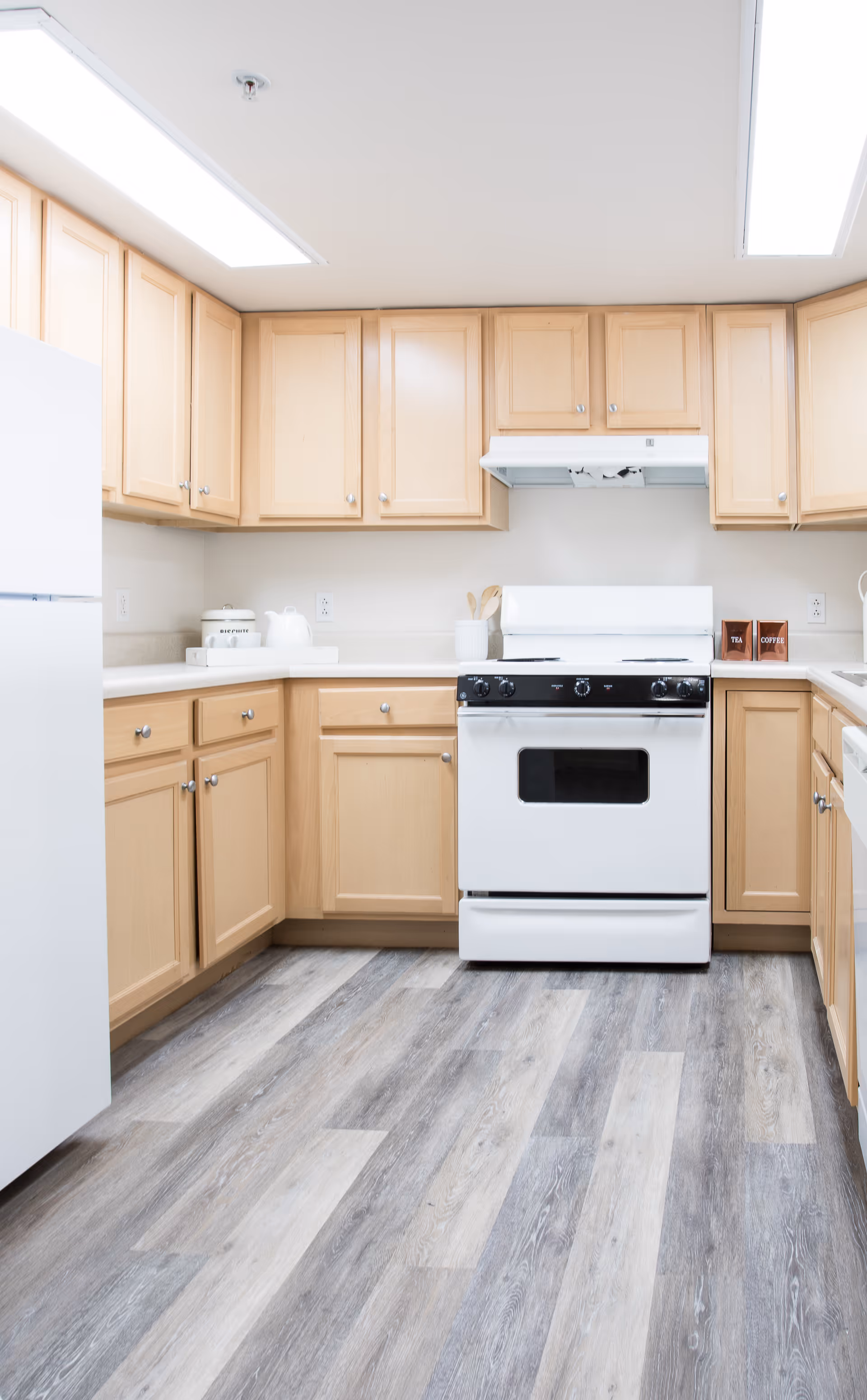 Bright kitchen with light wood cabinets, white countertops, a white refrigerator on the left, a white stove with a range hood in the center, and a light gray wood-patterned floor.