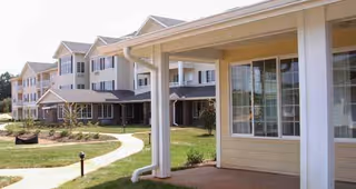 Exterior view of a multi-story retirement facility building with beige siding and white trim, surrounded by a well-maintained lawn and a curved concrete walkway.