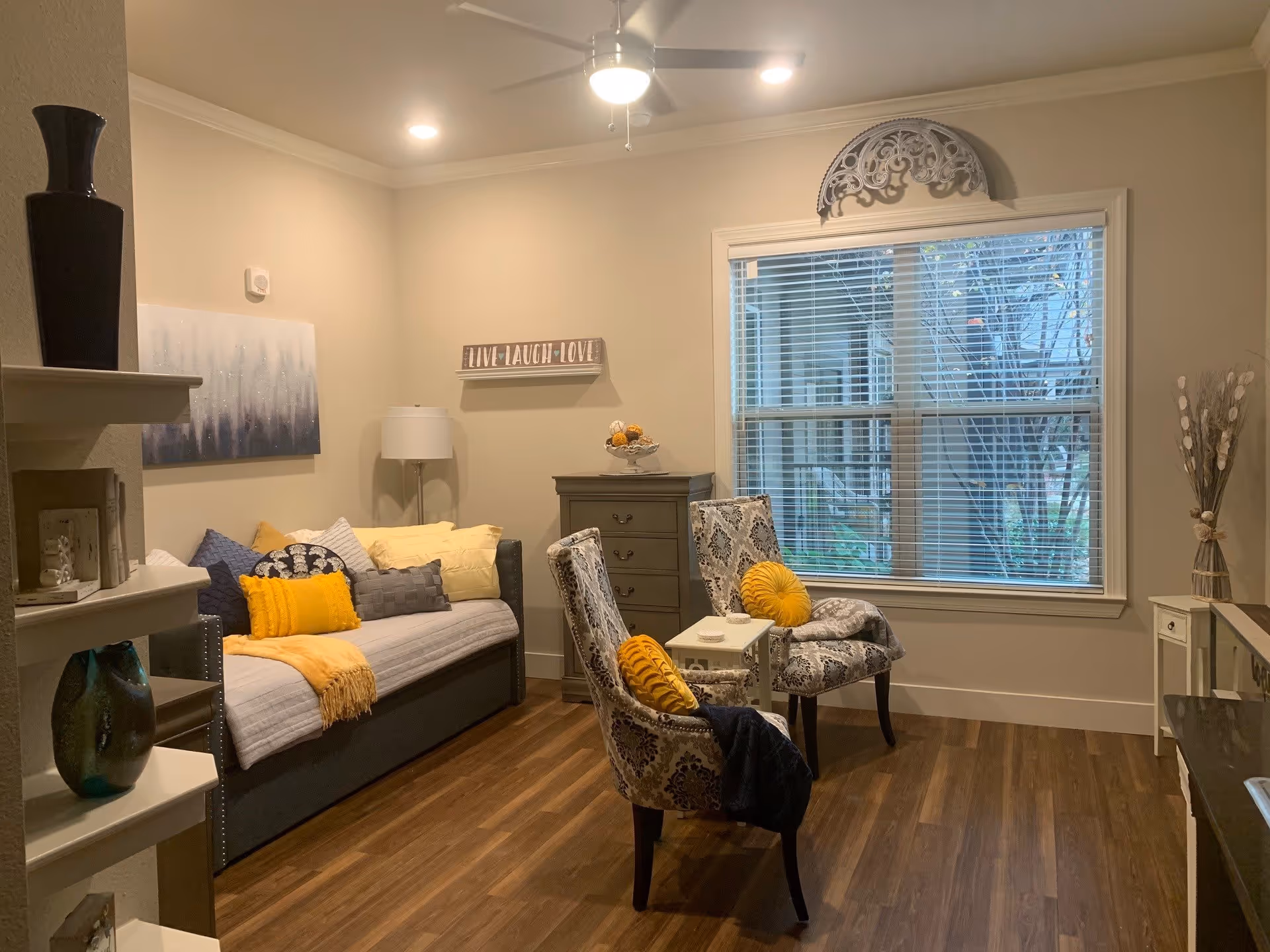 A cozy living room with a gray daybed adorned with yellow and gray pillows and a yellow throw blanket. Two patterned armchairs with yellow round pillows face a small white table. The room has wood flooring, a large window with blinds, a ceiling fan with lights, a tall lamp, a chest of drawers, and decorative wall art including a sign that reads 'LIVE LAUGH LOVE'.