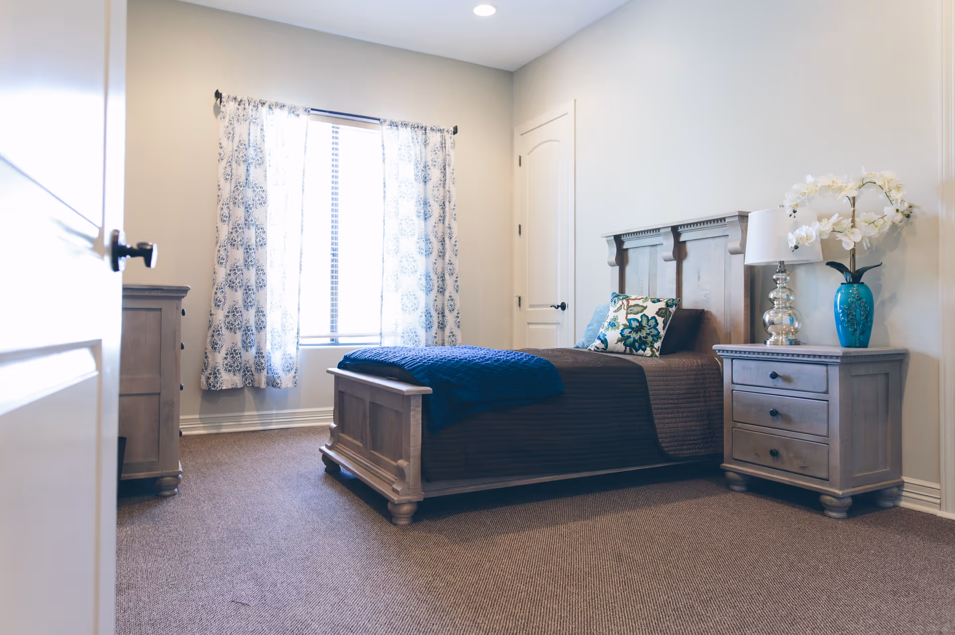 A bright bedroom with a wooden bed frame, a brown quilt, a blue throw blanket, and decorative pillows. There is a wooden nightstand with a lamp and a blue vase with white flowers. A window with patterned curtains lets in natural light, and a closed white door is visible on the right wall.