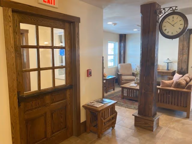 Interior view of a senior living facility common area with wooden furniture including a sofa, armchair, coffee table, and side tables. A large wooden pillar with a vintage-style clock is visible in the center. There is a wooden door with glass panes on the left and an exit sign above it.